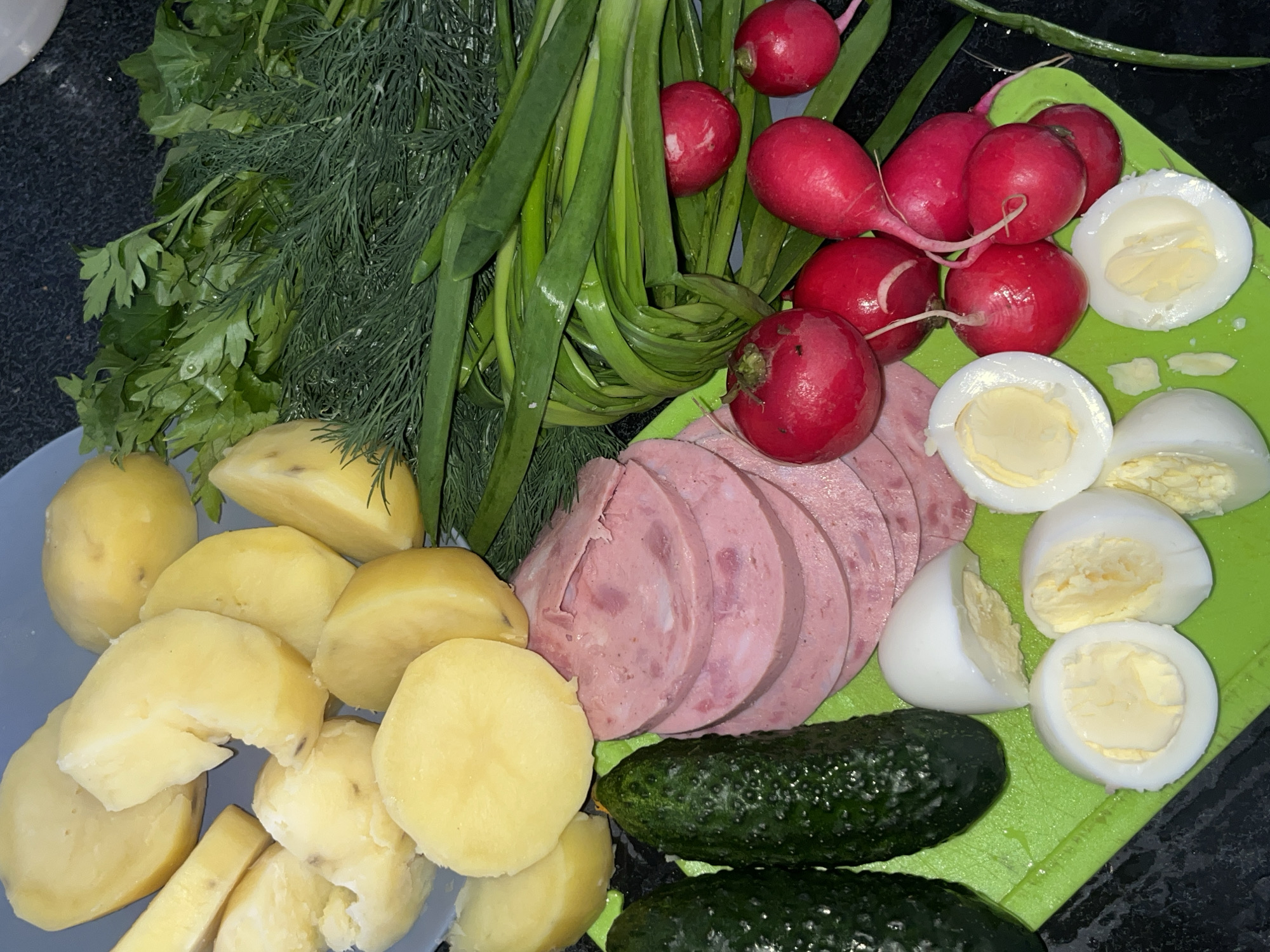 The image shows a selection of ingredients for a salad, likely okroshka, a cold soup popular in Russia. The ingredients are arranged on a dark surface and include: boiled potatoes, sliced sausage, chopped hard-boiled eggs, radishes, fresh dill, parsley, green onions, and cucumbers. All are on a gray table. The image was presumably taken in Krasnoyarsk, Russia.