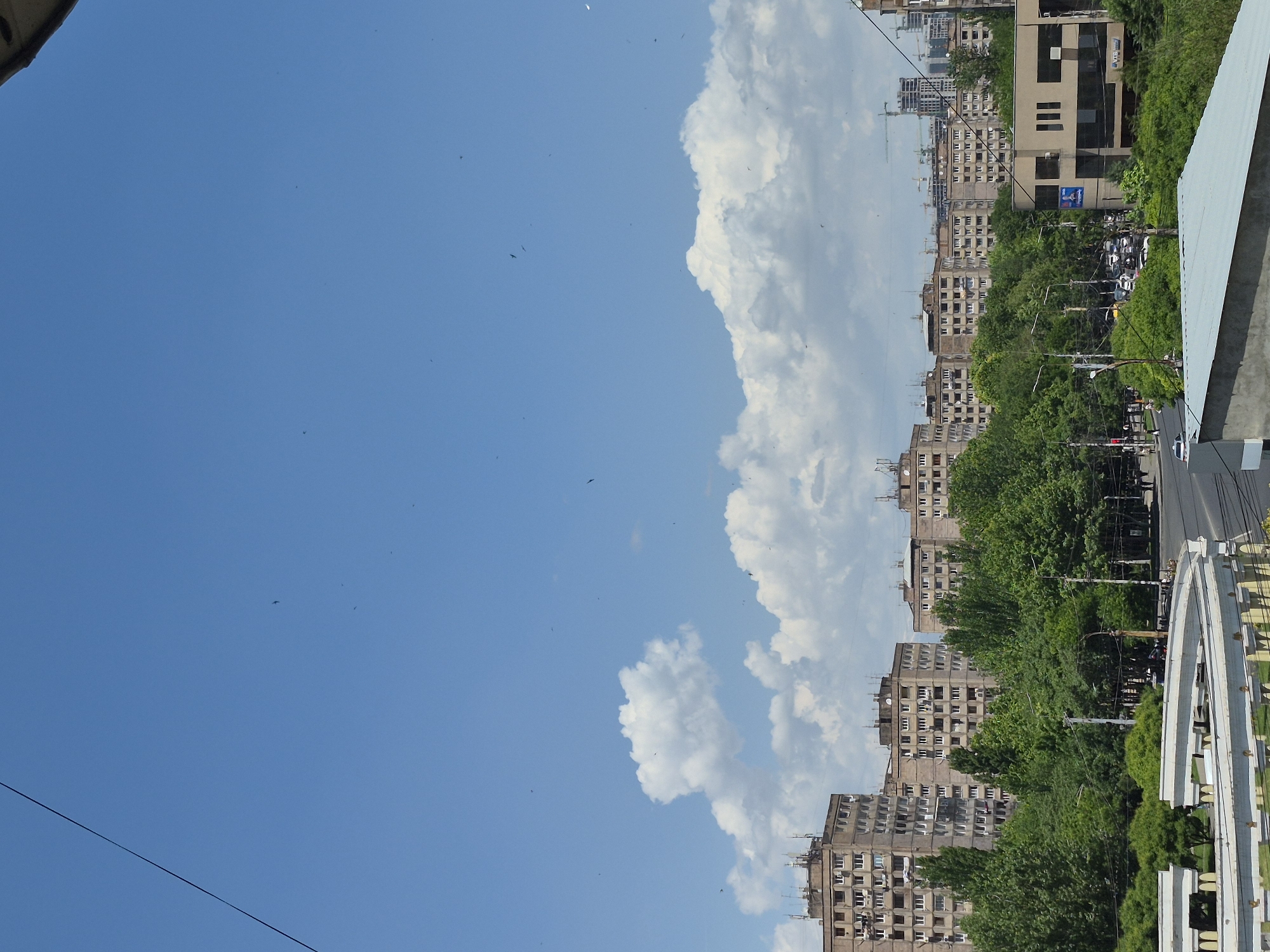 A wide, upright shot captures a vibrant cityscape in Yerevan, Armenia, under a clear blue sky during daylight hours. The top portion of the image is dominated by the expansive, bright blue sky, accented by a large, fluffy white cloud on the right side and several smaller clouds scattered across the frame. Numerous tiny dark specks, likely birds or insects, are visible against the sky.
Below the horizon, the city unfolds with rows of multi-story apartment buildings, primarily light brown or beige, featuring many windows and some rooftop satellite dishes. These buildings line streets that are heavily populated with lush, green trees, whose dense canopies partially obscure the lower portions of the structures. In the distant background, several construction cranes indicate ongoing urban development.
In the mid-ground, a more modern building with dark, reflective windows and light-colored panels stands out. A blue sign, possibly an advertisement or company logo, is visible on its facade, though the text is illegible. Utility poles and an intricate network of overhead wires crisscross the frame, particularly noticeable against the sky and above the buildings. A road with visible vehicles runs between the rows of trees and buildings.
To the bottom right, a distinctive white architectural structure with several arched elements, possibly a bridge or monument, adds a prominent feature to the foreground. The scene conveys a sense of a bustling city on a bright, sunny day, characterized by a blend of urban infrastructure and abundant greenery.