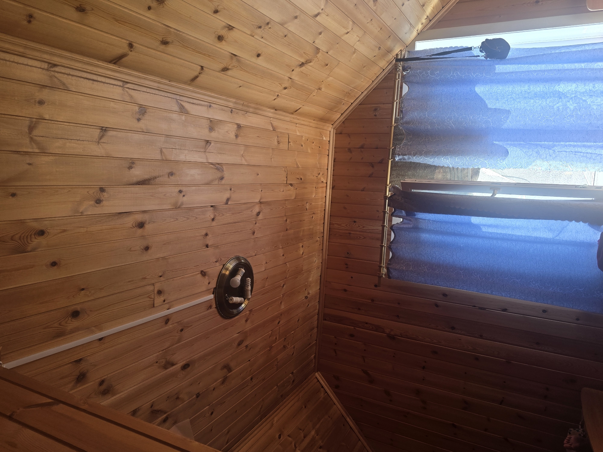 The image depicts the interior of a room with a wooden paneled ceiling and walls. In the upper part of the image, a circular light fixture with several bulbs is attached to the ceiling. The ceiling planks run diagonally from left to right and upwards. The walls are also paneled with wood and are darker in color than the ceiling. Two blue curtains hang across a window, partially obscuring the view outside. A strap with a black bag hangs from the curtain rod on the right side of the window. The room appears to be located in Maksimiha, Russia.