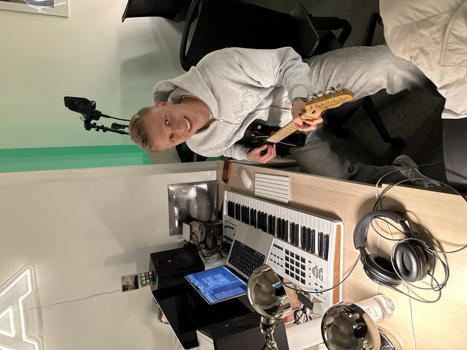 A fair-skinned man in a gray hoodie and sweatpants sits in a black chair, smiling and holding a black electric guitar. A music production setup includes a keyboard, computer screen with audio software displayed, speakers, headphones, and a microphone on a stand. Additional items on the desk are a water bottle and a Rubik's cube. A neon A is visible on the wall.