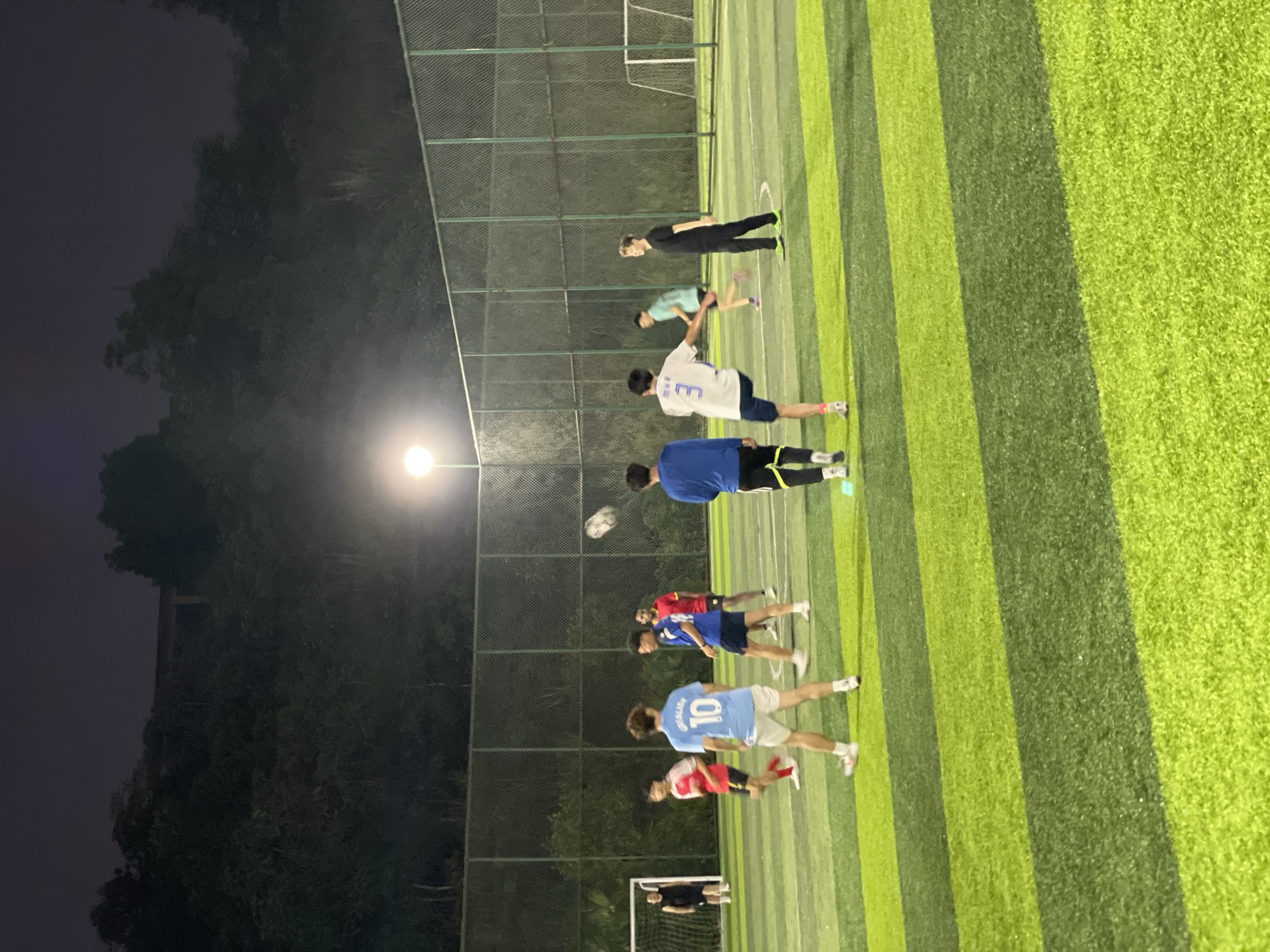 Captured at night in Jiangjin, China, the scene presents a soccer match on an artificial turf field with striped green and light-green patterns. Players wearing a variety of colored jerseys are positioned along the field. A bright overhead light illuminates the field, while a dark sky and trees form the background. A perimeter fence surrounds the field.