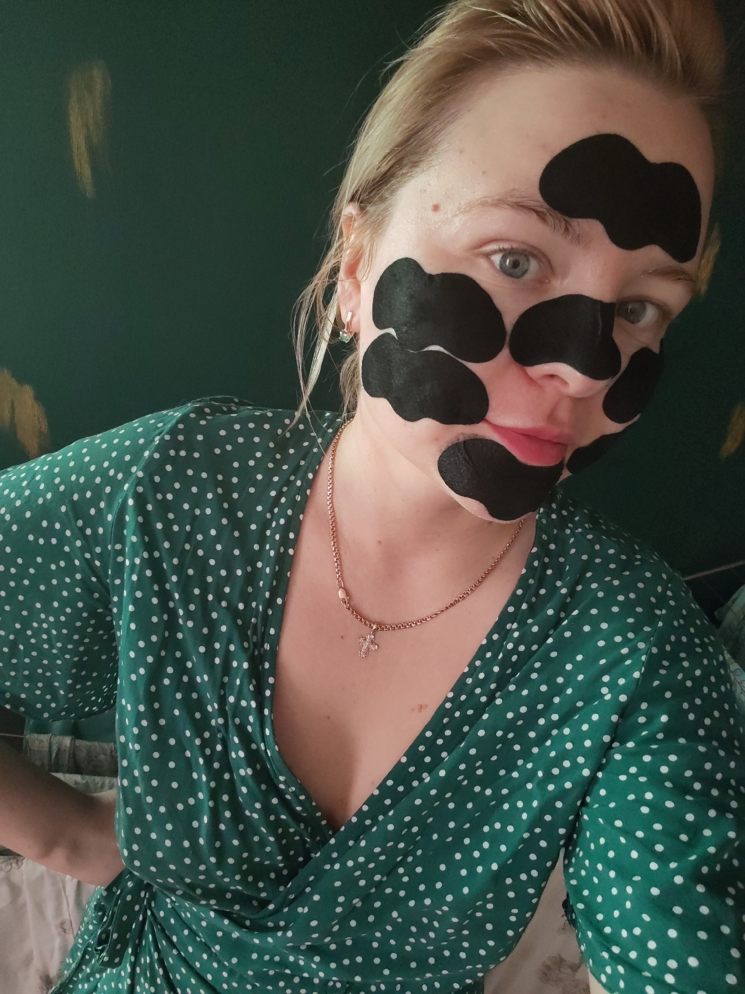 A blonde woman with light blue eyes, wearing a green polka dot wrap dress and a rose gold necklace with a cross pendant, is positioned in the foreground of the image. Her face is partially covered with black pore strips. She is looking directly at the camera. The background consists of a dark green wall with a golden abstract design on the upper left side. The lighting suggests an indoor setting. The image was taken in Izhevsk, Russia.