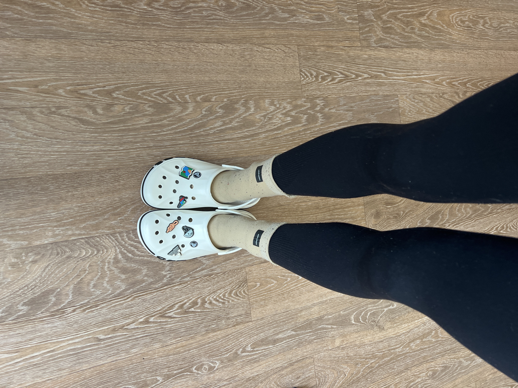 A pair of legs wearing black leggings and beige socks with black cuffs are positioned on a wooden floor. The feet are shod in white Crocs adorned with charms. The location appears to be indoors, likely in a residential or commercial space featuring wood-patterned flooring. The image does not reveal any specific information about the city of Chaikovsky, Russia.