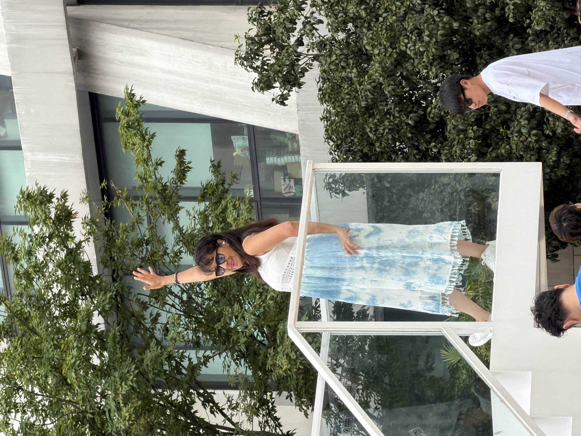 A medium shot captures a woman standing on a white staircase with a glass railing, waving with her right hand. She is wearing a white sleeveless top with a crochet detail and a long, blue tie-dye skirt with a fringed hem. She has dark hair and is wearing sunglasses and white earrings. To the right of the staircase, a young man in a white t-shirt and black sunglasses is visible from the chest up, looking to his left. Behind the subjects, lush green trees obscure a modern building with large windows. The overall scene suggests an outdoor setting in an urban environment.