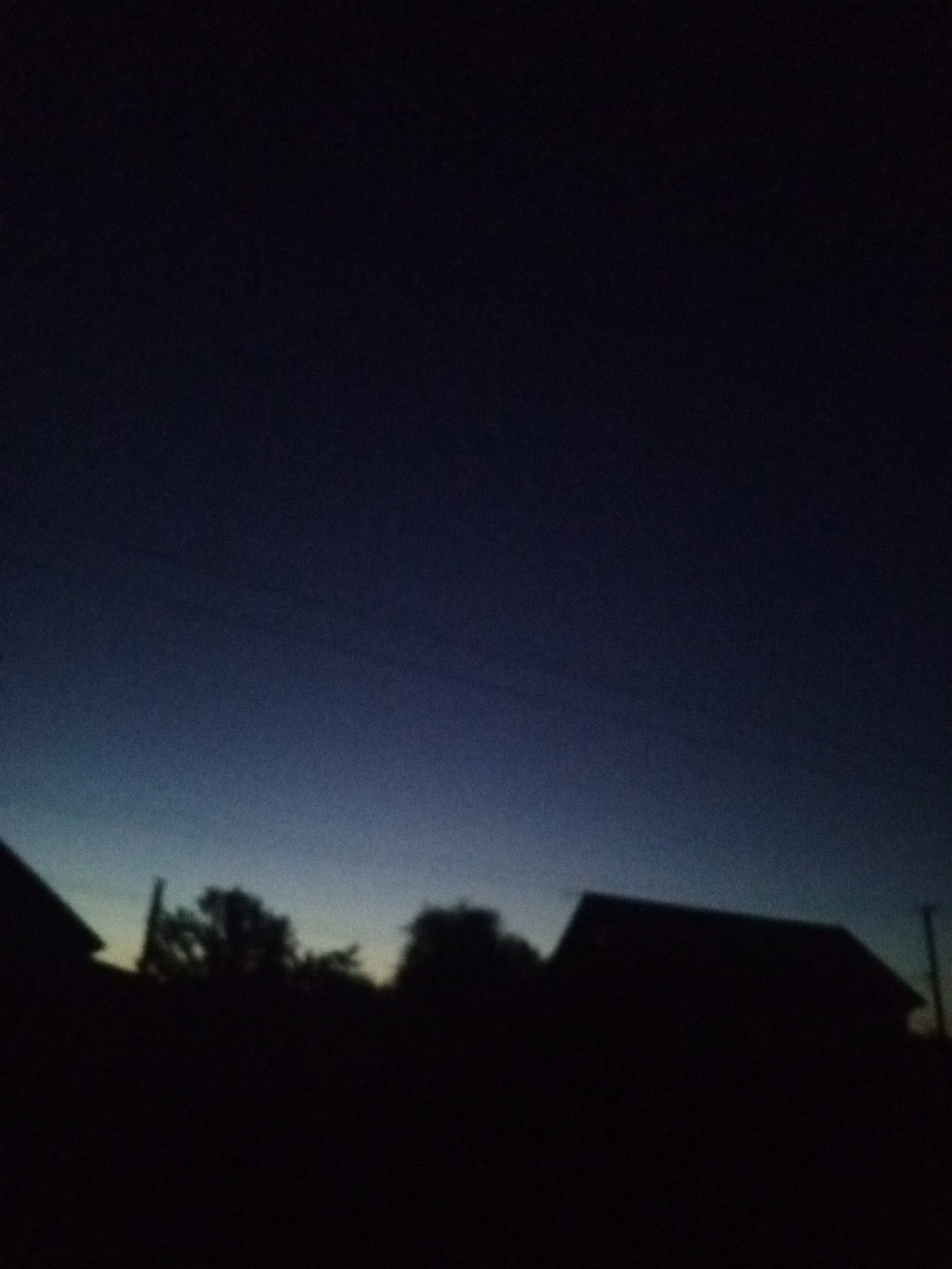 The image captures a silhouetted landscape against a dark, twilight sky. In the foreground, the dark outlines of several buildings and trees are visible. One building on the right has a prominent gabled roof. The sky transitions from a deep, dark blue at the top to a lighter, almost teal hue near the horizon. Faint horizontal lines, possibly power lines, are discernible against the darker parts of the sky. The scene appears to be set in a rural or suburban area, given the presence of houses and trees. The location is identified as Buraly, Russia, suggesting a view of dwellings and natural elements within that locale during dusk or dawn.