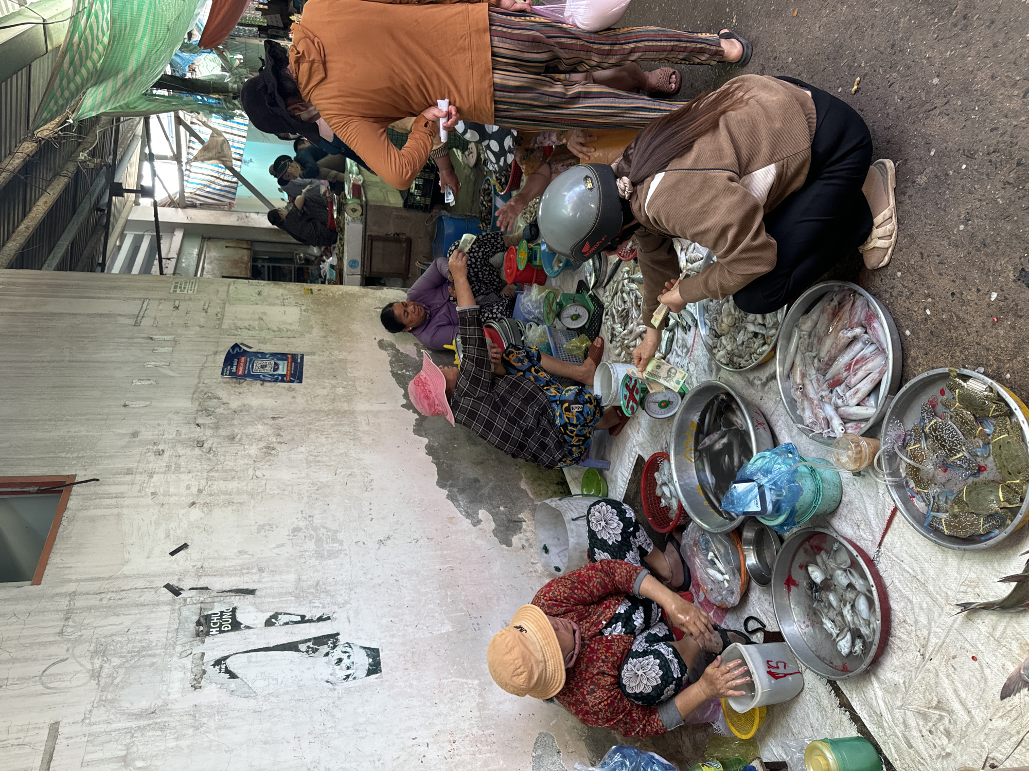 The scene is a busy, informal outdoor fish market in Mũi Né, Vietnam. Several vendors sit on the ground surrounded by metal bowls and buckets filled with various types of seafood including shrimp, squid, and crabs. A customer wearing a helmet and brown jacket is kneeling and paying a vendor with Vietnamese Dong banknotes. Other people are present in the background and around the vendors. The ground is a combination of pavement and a white tarp. A concrete wall with old posters partially torn off is visible to the left. Overhead, there is a patchwork of shade covers.