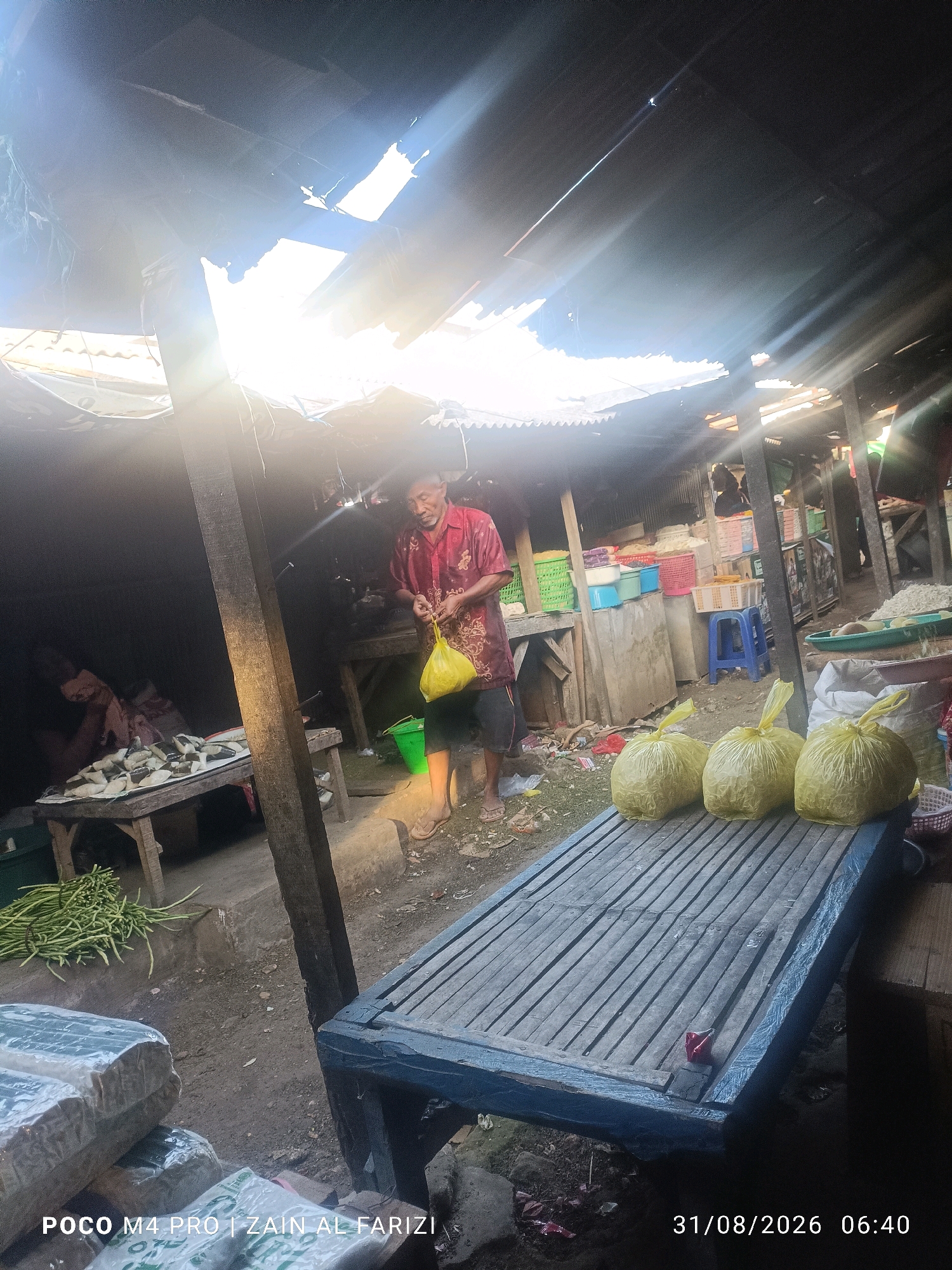 In a market stall in Arosbaya, Indonesia, sunlight streams through a damaged roof, creating lens flares and illuminating a vendor. The vendor, an adult male wearing a red patterned shirt and black shorts, stands near a table laden with fish. He holds a yellow plastic bag in his right hand and appears to be in the process of selling or displaying goods. In the foreground, three yellow plastic bags are arranged on a wooden surface. The market environment is rustic, with wooden structures and various baskets and containers visible in the background, suggesting a bustling commercial setting. The ground appears to be dirt and debris. The overall impression is one of a working market, likely operating in the early morning given the timestamp of 06:40.