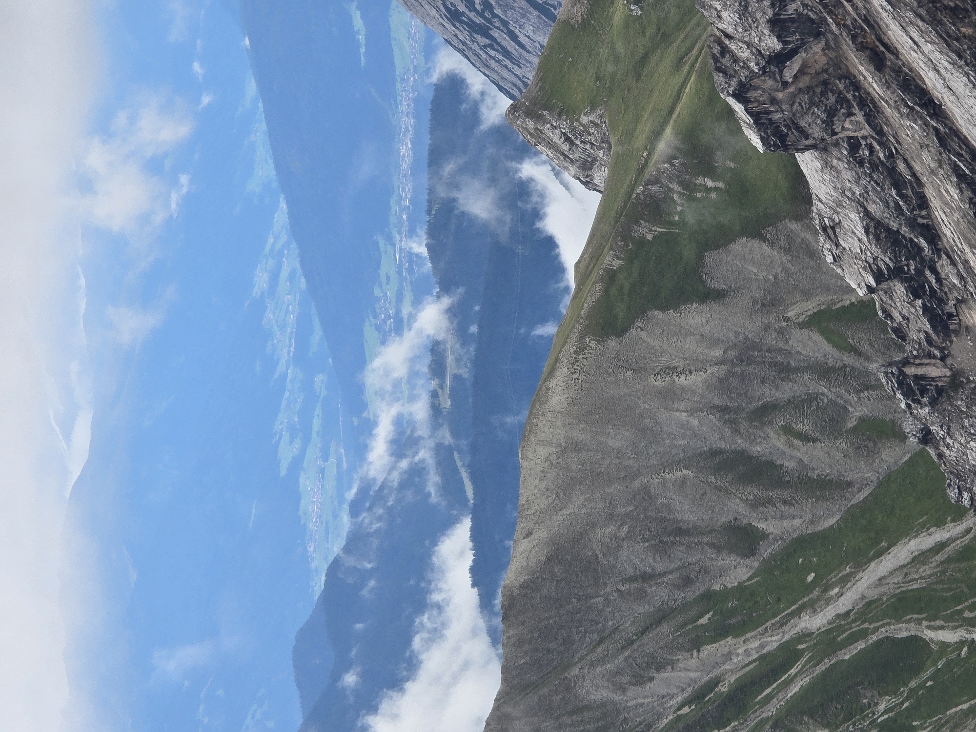 This is a vertical, eye-level, medium shot photograph taken from a high vantage point overlooking a mountain range. The foreground is dominated by a steep, rocky, and grassy slope with distinct textural variations. Moving into the midground, there are more layered mountain ranges, some of which are partially obscured by clouds. The background features a hazy, light blue sky with further layers of distant mountains and a small village nestled in a valley. The overall impression is one of vastness and natural beauty. The scene is located in the vicinity of Garmisch-Partenkirchen, Germany, a town renowned for its Alpine scenery.