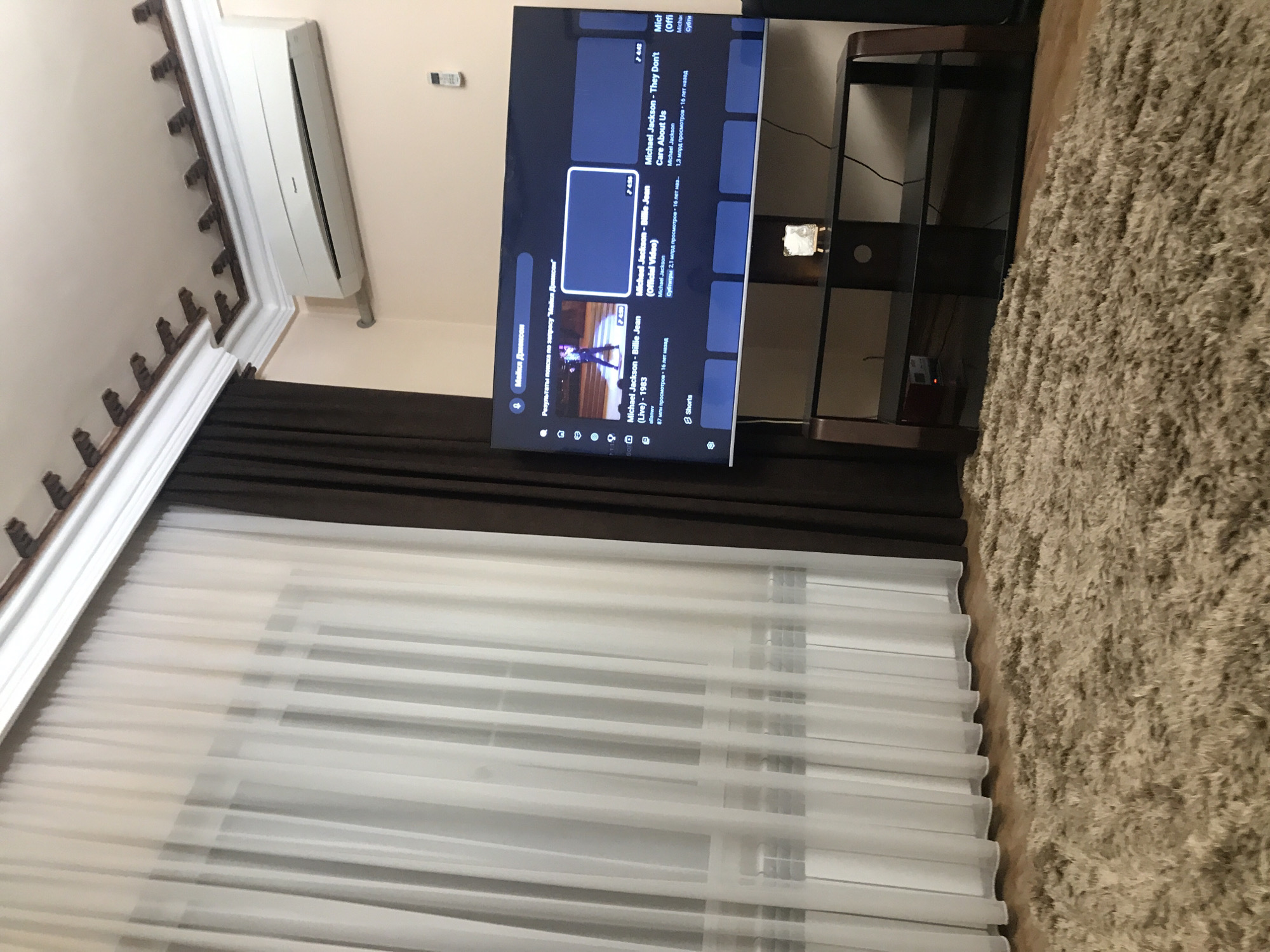 A residential interior features a television displaying a YouTube interface with search results for "Michael Jackson," including videos like "Billie Jean" and "They Don't Care About Us." The TV is placed on a dark-colored TV stand. Adjacent to the TV stand is a shag rug, and window curtains with a sheer layer partially obscure daylight. An air conditioner is visible mounted on the upper wall. The location is assumed to be in Yerevan, Armenia.