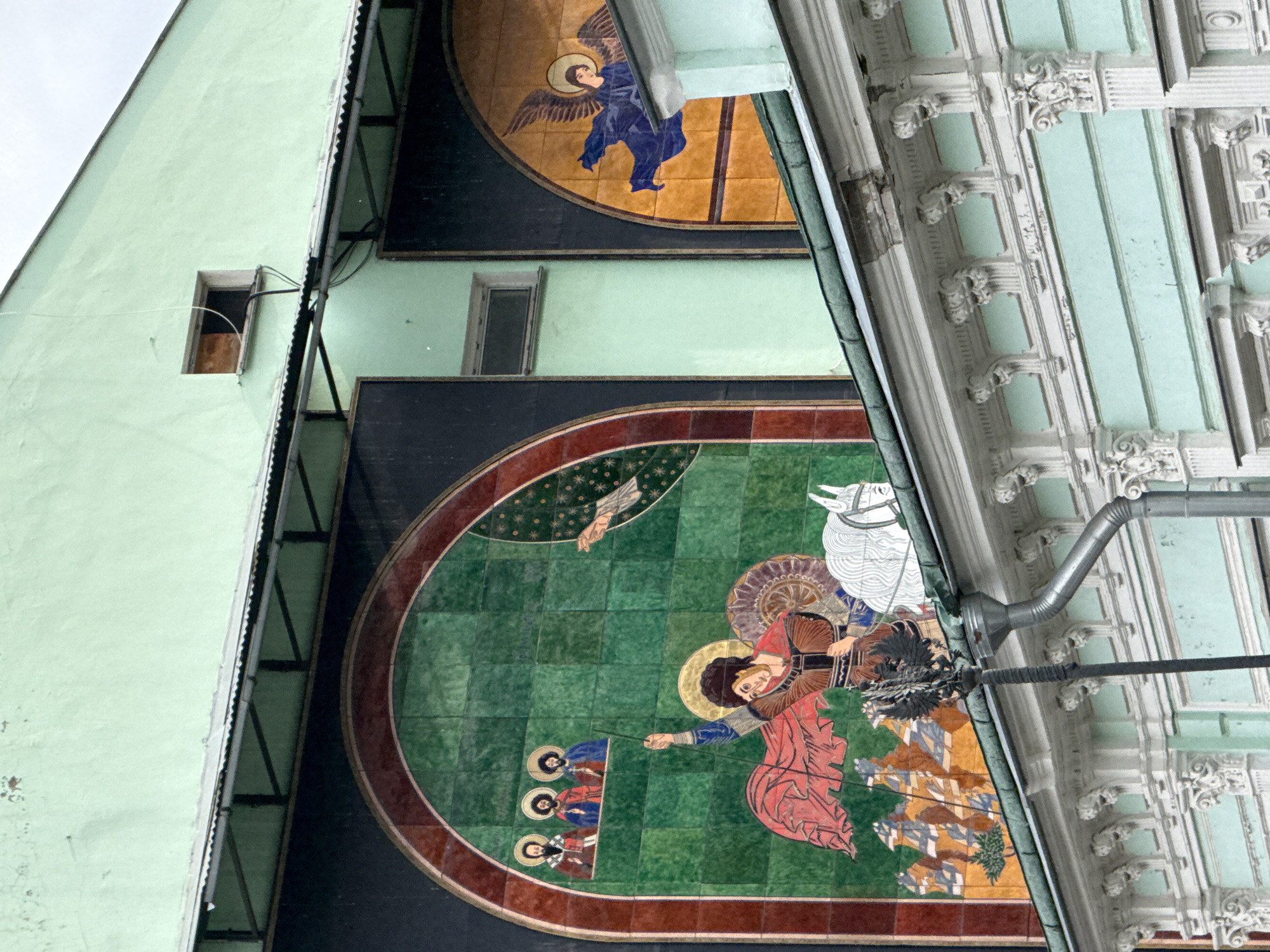 The image depicts a section of an exterior wall of a building, likely in Moscow, Russia, adorned with intricate mosaics. The mosaics feature religious or historical figures, rendered in vibrant colors.
In the upper part of the image, a mosaic of an angel with blue robes and wings is visible against a yellowish background. Below this, on a light green painted section of the building, a small, open window is present. Further down, a larger, curved mosaic dominates the scene. This mosaic is set against a dark background and features a central figure, possibly an angel or a saint, with a halo and reddish robes, pointing a staff towards a group of three figures in blue robes. To the right of the central figure, a white horse is depicted. The background of this mosaic is a vibrant green, with star-like patterns and a red border.
The building itself has a light green facade on the left, and on the right, there's a section of ornate architectural detailing in a pale blue and white color scheme, featuring columns and decorative elements. A metal downspout is visible running across the upper right portion of the image. The sky appears overcast, suggesting a cloudy day. The overall impression is of historical and artistic decoration on an older building.