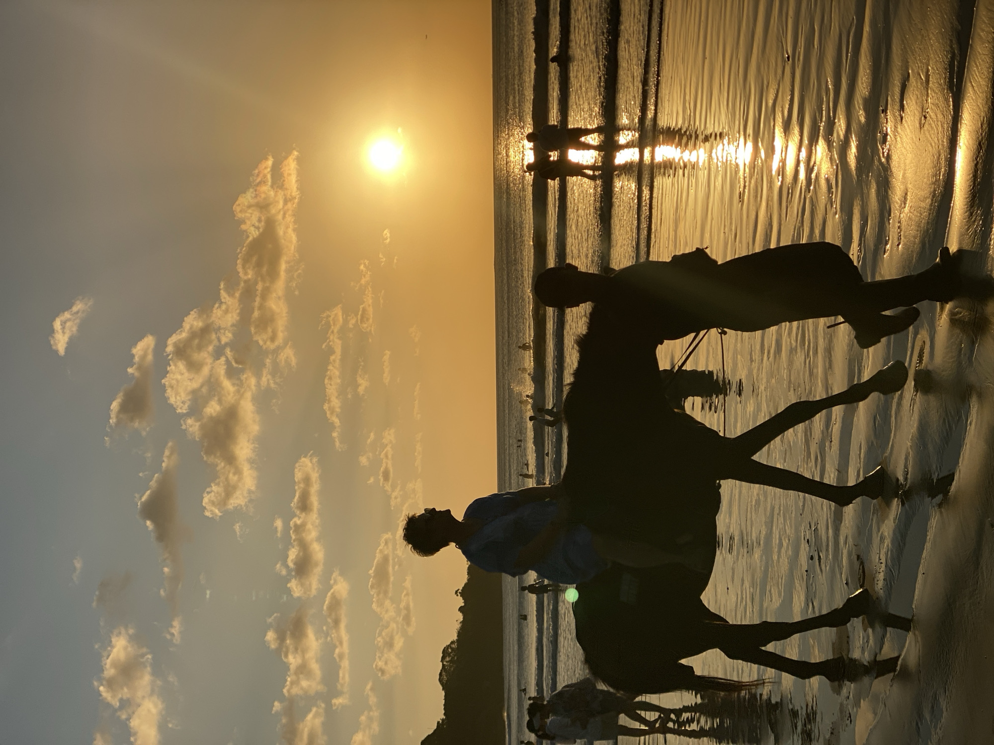 A silhouette of a woman riding a horse along the beach in Kuta Selatan, Indonesia, is captured at sunset. The horse is being led by a person whose silhouette is also visible. The sun is setting over the ocean, casting a warm golden glow on the scene and reflecting on the wet sand. Several other people are visible in the background, wading in the shallow water and walking along the shoreline. The sky is filled with scattered clouds illuminated by the setting sun. The overall atmosphere is serene and picturesque.