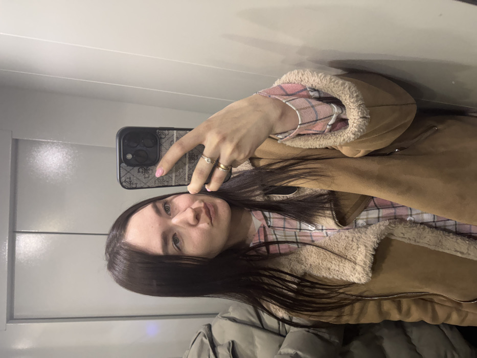 The image displays a young adult female taking a selfie in an indoor setting, likely an elevator or narrow hallway, in Chaikovsky, Russia. The female has long, dark brown hair and light eyes, looking slightly to her right with pursed lips. She is dressed in a light brown faux suede jacket with cream-colored shearling lining, worn over a pink and light-colored plaid shirt. Her left hand holds a smartphone, encased in a patterned black case, positioned to capture her reflection. Her fingernails are painted pink, and she wears two rings on her left ring finger. A partial view of an indistinct individual or object, wearing a light-colored padded jacket, is visible on the far left. The background consists of smooth, light-colored walls with visible vertical seams.