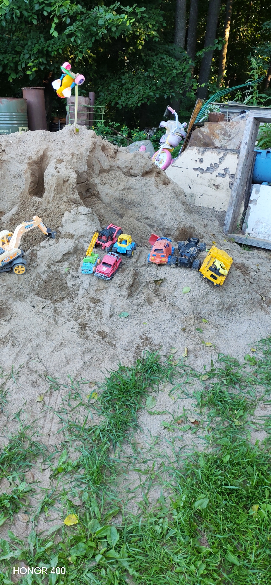 A sandbox filled with various toy vehicles, including a yellow excavator, a red car, a blue truck, an orange truck, a black truck, and a yellow dump truck, is situated outdoors amidst lush green foliage. In the background, a large sand mound is present, adorned with a colorful pinwheel on a stick. To the right, a section of a white structure with peeling paint is visible, along with a blue barrel. The scene appears to be in a residential area in Noviy, Russia, given the presence of the sandbox and playground-like elements.