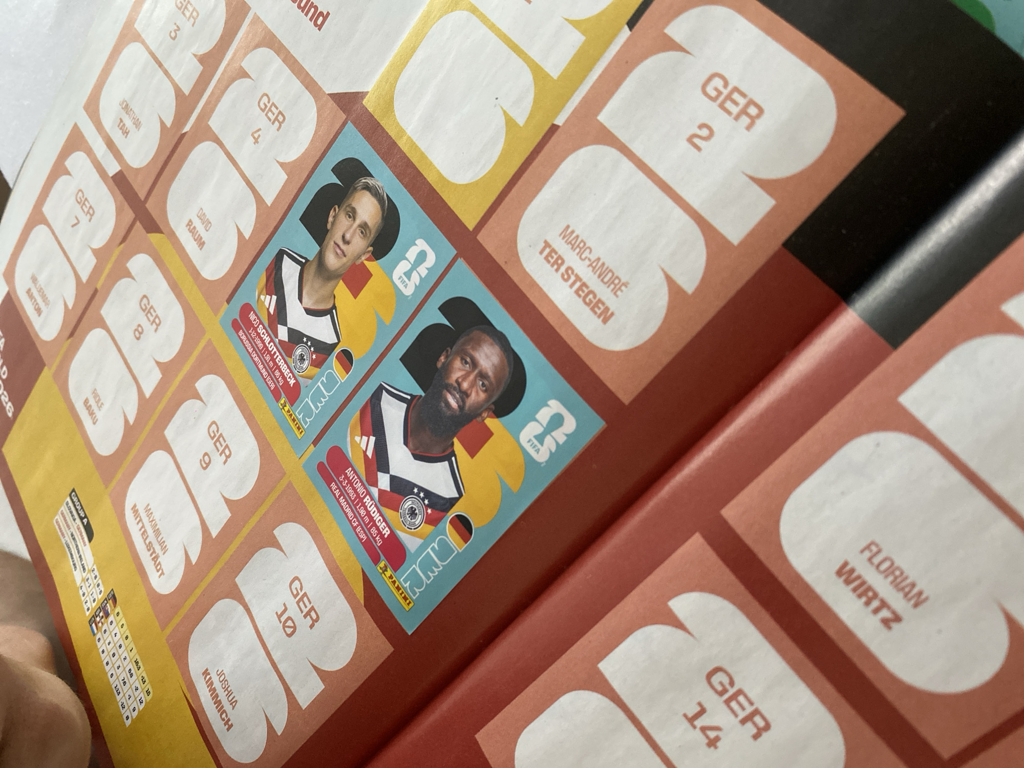 The image shows a partially filled Panini FIFA World Cup 2026 sticker album. Visible are several German national team player stickers and empty slots for additional stickers. The stickers include Nico Schlotterbeck, Antonio Rüdiger, and Marc-André ter Stegen. Empty slots are labeled with "GER" and numbers 2, 3, 4, 7, 8, 9, 10, and 14, along with the player's names that correspond to those stickers. A small section showing group A match results and a calendar are also visible.