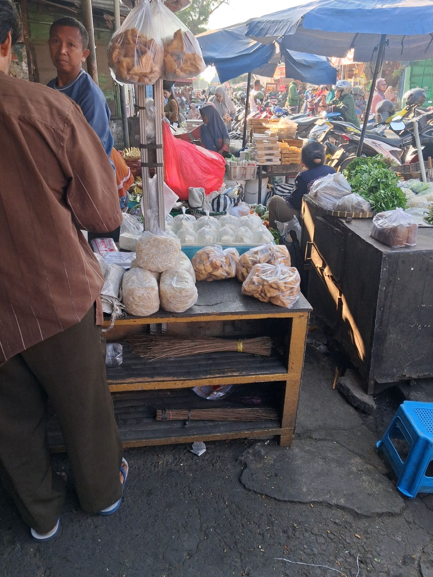 A busy outdoor market in Surakarta, Indonesia, features a stall with various food items for sale. In the foreground, a person wearing a brown long-sleeved shirt and dark pants stands with their back to the camera, facing the stall. Another person, a man with short dark hair, is partially visible on the left, looking towards the viewer.
The stall itself is constructed from wood and metal, with shelves displaying packaged food items. Prominently featured are several clear plastic bags filled with small, dried shrimp, and other bags containing fried snacks, possibly crackers or pastries. Brooms are also stacked on a lower shelf. Behind the stall, a variety of other market activities are taking place. People are seen milling about, some wearing headscarves, and several motorcycles are parked, suggesting transportation for both vendors and customers. Blue and white tarps are stretched overhead, providing shade. The ground is paved with asphalt, showing signs of wear and tear. A bright blue plastic stool is visible to the right of the frame. The overall atmosphere is one of bustling commerce under a bright sky.