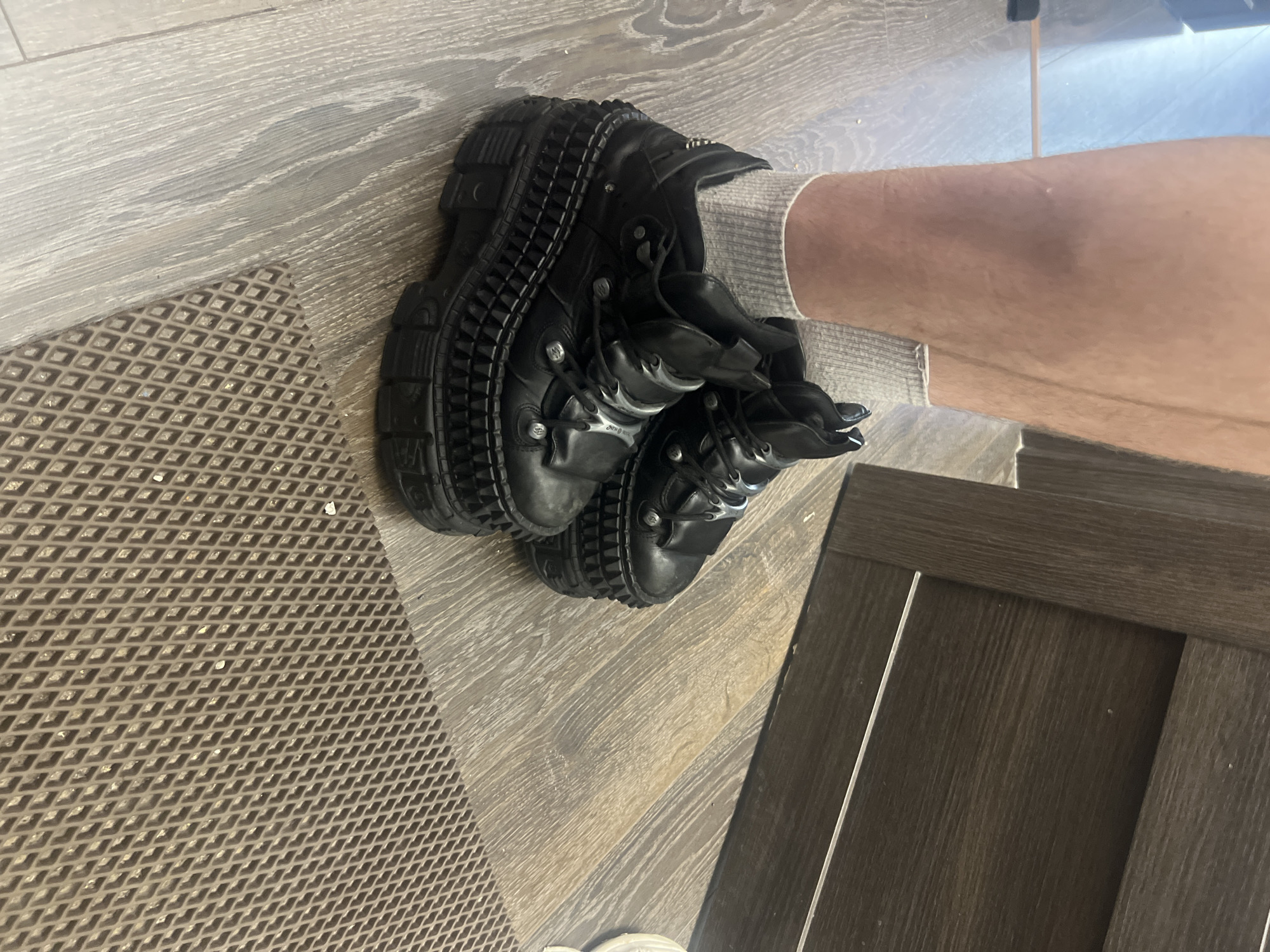 An angled, eye-level indoor shot shows a person's lower leg and foot, shod in a striking black boot and light grey sock. The setting, identified as Saint Petersburg, Russia, features a light-colored wooden floor with a visible grain, where the foot rests. Part of a textured brown mat with a diamond grid pattern is also visible, extending from the bottom left. A dark brown wooden surface, possibly furniture or a door, is seen in the bottom right.
The central subject is a robust, black platform boot, characteristic of the New Rock brand, though the text is partially obscured; "New Rock" or a stylized version is faintly visible on the shoe's side near the lacing. The boot boasts an aggressive, multi-layered lug sole with prominent ridges and spikes, giving it a heavy-duty, industrial appearance. The upper part of the boot is embellished with various metallic, silver-toned accents, including small studs and decorative flame-like or wing-like elements. The person wears a light grey, ribbed sock, which extends above the boot and below the bare lower leg. The lighting is soft and ambient, typical of an indoor environment, making the time of day indeterminate. No specific activity is captured other than the foot resting on the floor, and no emotions are conveyed.