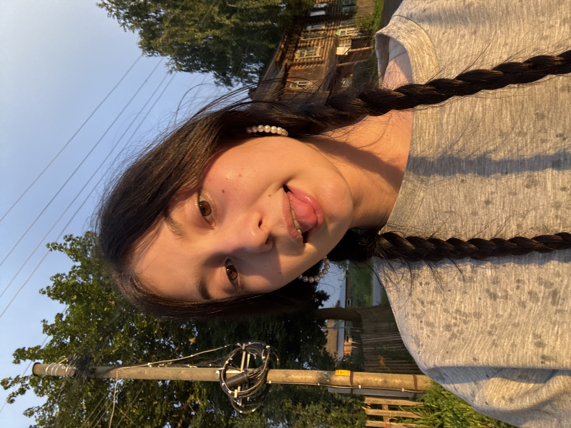 A young woman with dark hair in pigtails sticks her tongue out for a selfie outdoors. She wears a grey t-shirt and large pearl hoop earrings. Behind her, a rustic wooden house and a utility pole are visible under a clear blue sky. The lighting suggests it is late afternoon or early evening.