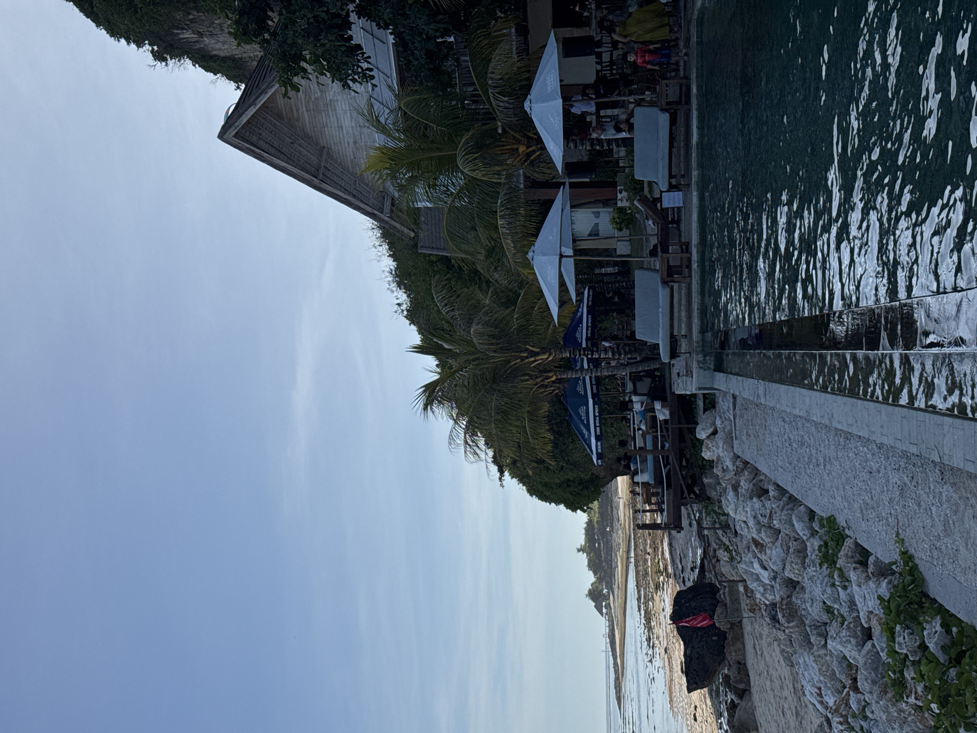 The image shows a seaside scene in Kuta Selatan, Indonesia. A building with a wooden roof sits atop a cliff, partially obscured by palm trees and other vegetation. Below the cliff, there is a beach, with visible rocks, sand, and a receding tide. A stone retaining wall separates the beach from a pool deck area where tables and chairs are set up under white umbrellas. The sea stretches out towards the horizon, covered by a clear and pale blue sky. The lighting suggests it is daytime, possibly the late morning or early afternoon.
