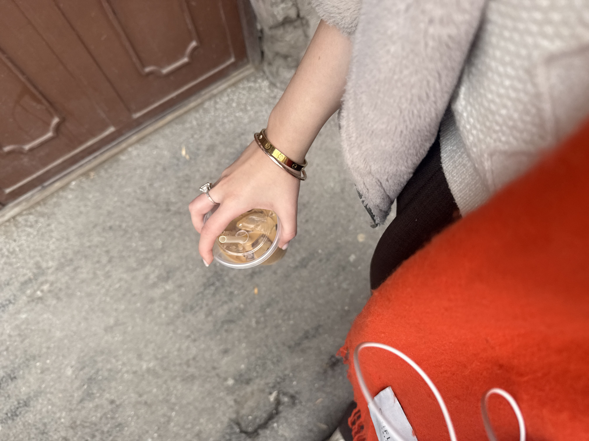 An overhead, close-up photograph depicts a person's left arm and upper torso in an outdoor setting within Yerevan, Armenia. The fair-skinned left hand, adorned with a silver-toned ring featuring a prominent square-cut gemstone on the ring finger, securely grips a transparent plastic cup. The cup contains a light brown iced beverage with ice cubes and a white straw. Two bracelets are visible on the wrist: a gold-toned bangle with recessed screw-head details and a thinner, plain silver-toned bangle. The person is dressed in layered clothing, including a light grey, fuzzy or plush outer garment and a section of bright orange, textured fabric with white decorative stitching and a white label. The ground below is a mottled grey, rough-textured surface, consistent with concrete or pavement. In the upper right background, a vertical section of a dark brown wooden door or panel, featuring decorative raised molding, is visible.