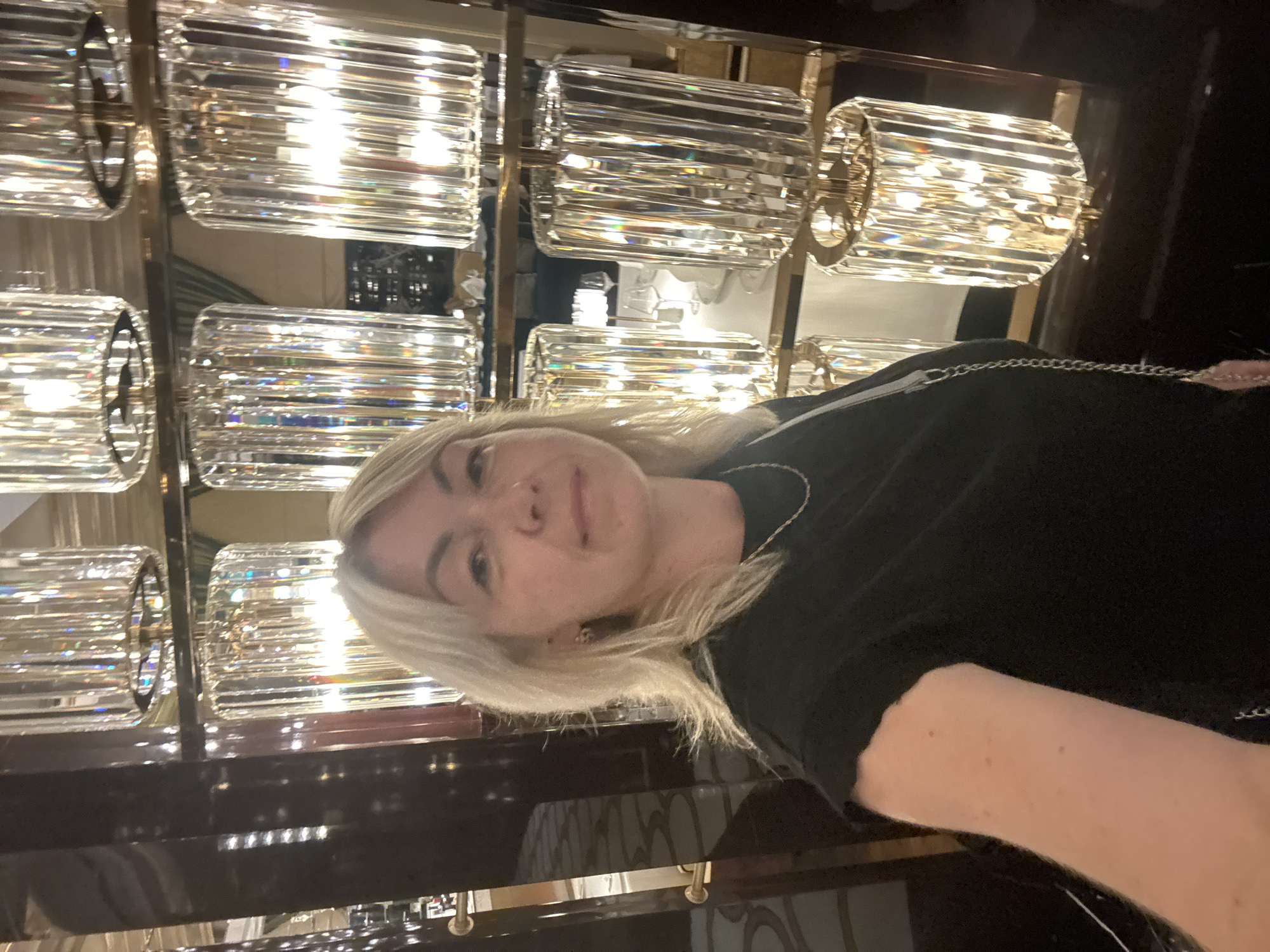 A medium shot, selfie-style image shows a blonde woman with fair skin and a light dusting of freckles on her arm. She is wearing a black t-shirt and a thin, silver chain necklace. Her hair is shoulder-length and styled with a side part. The background is dominated by rows of tall, cylindrical crystal chandeliers with a warm, internal glow. These are arranged on shelves against a dark, reflective surface, possibly a wall or furniture. The lighting is bright and creates numerous reflections and refractions off the crystal. The environment appears to be an interior space, likely a hotel lobby or a high-end establishment, with an opulent and decorative aesthetic. Based on available information, this scene is consistent with the interior of a luxury hotel in Estosadok, Russia, possibly the Radisson Blu Resort & Congress Centre or similar establishment known for its elaborate decor.