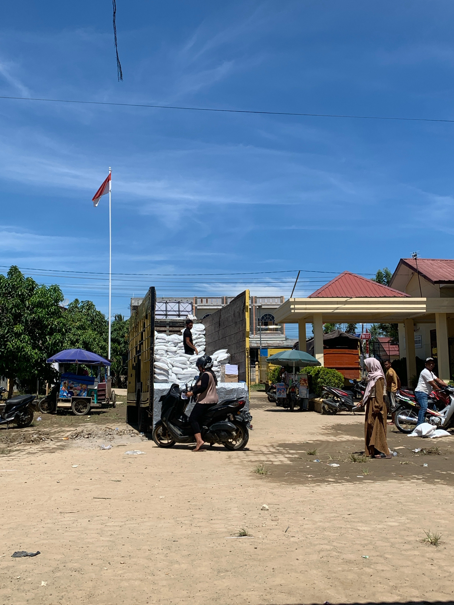 The image depicts a daytime scene in what is identified as Pantegurah, Indonesia, characterized by a dry, dusty ground and a clear blue sky with some wispy clouds. A tall flagpole with the Indonesian flag is prominently displayed. In the foreground, a person is seated on a black scooter, wearing a brown top and dark pants. To the left of the scooter, a food cart with a blue umbrella is parked, displaying colorful pictures. Behind the scooter, a large truck is parked, loaded with many white sacks, possibly containing rice or similar goods. Two individuals are visible near the truck, one standing on the truck bed with a white sack on his head, and another partially obscured. Further in the background, a building with a red roof and cream-colored walls is visible, along with other structures and parked motorcycles. Several other people are also present, some walking and others on motorcycles. The environment appears to be a commercial or communal area, possibly a market or a transport hub, given the presence of goods, vehicles, and food vendors. The overall impression is one of a busy, active day in a tropical climate.