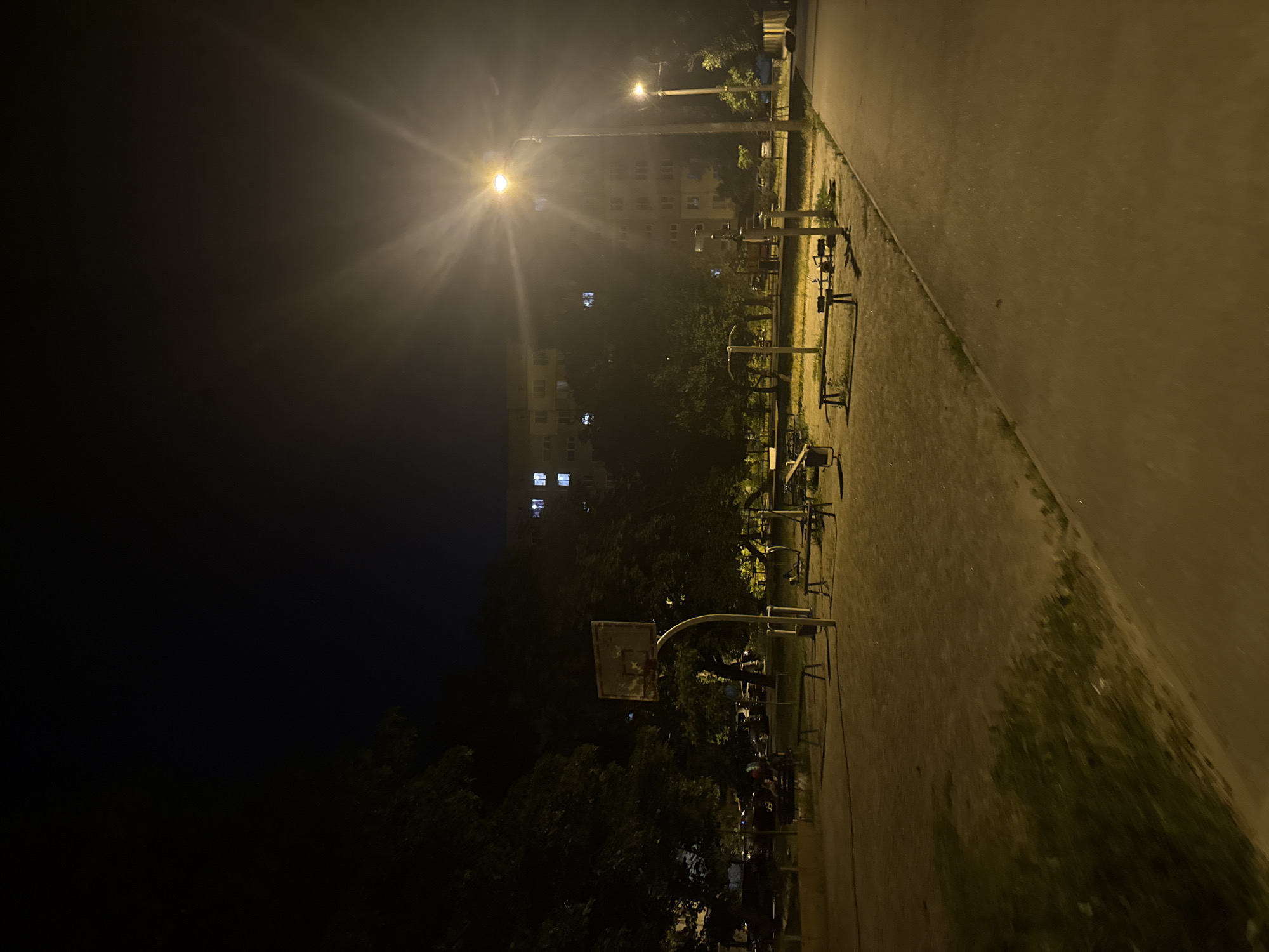 This image depicts an outdoor basketball court and a small outdoor exercise area at night in the city of Dnipro, Ukraine. A bright street lamp illuminates the scene, casting long shadows. In the foreground, a paved walkway curves to the right. To the left, the basketball court is visible with a hoop and backboard. Beyond the court, an exercise area features several pieces of equipment, including benches and pull-up bars. A multi-story apartment building with lit windows is visible in the background, partially obscured by trees. Some vehicles and indistinct figures can be seen in the distance, suggesting the area is a public space within a residential neighborhood.