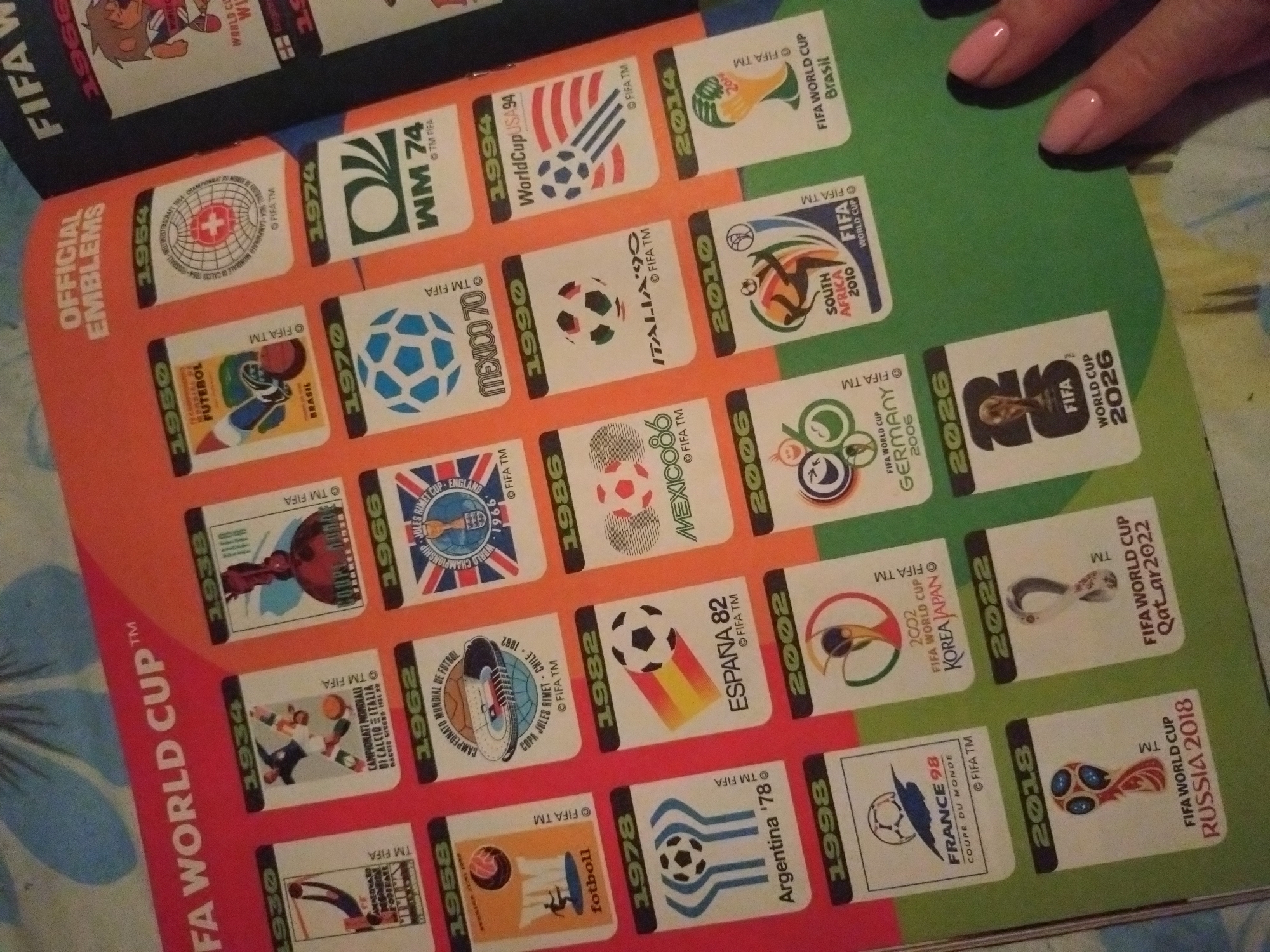 The image is a close-up photograph of a page from a sticker album, likely about FIFA World Cup emblems. The page is mostly a bright orange with a green border at the bottom. Various rectangular stickers are arranged in a grid pattern, each depicting the official emblem and year of a different FIFA World Cup.
The stickers visible represent the following years: 1930, 1934, 1938, 1950, 1954, 1958, 1962, 1966, 1970, 1974, 1978, 1982, 1986, 1990, 1994, 1998, 2002, 2006, 2010, 2014, 2018, 2022, and 2026. Each sticker contains the emblem, the country that hosted the World Cup, and "FIFA TM" or "© FIFA TM".
In the bottom right corner, a person's hand with light pink painted fingernails is visible, obscuring part of the green border and the 2026 sticker. The image is taken from a slightly elevated angle, looking down onto the page.
There is no information in the image to suggest a location in Stara Zagora, Bulgaria.