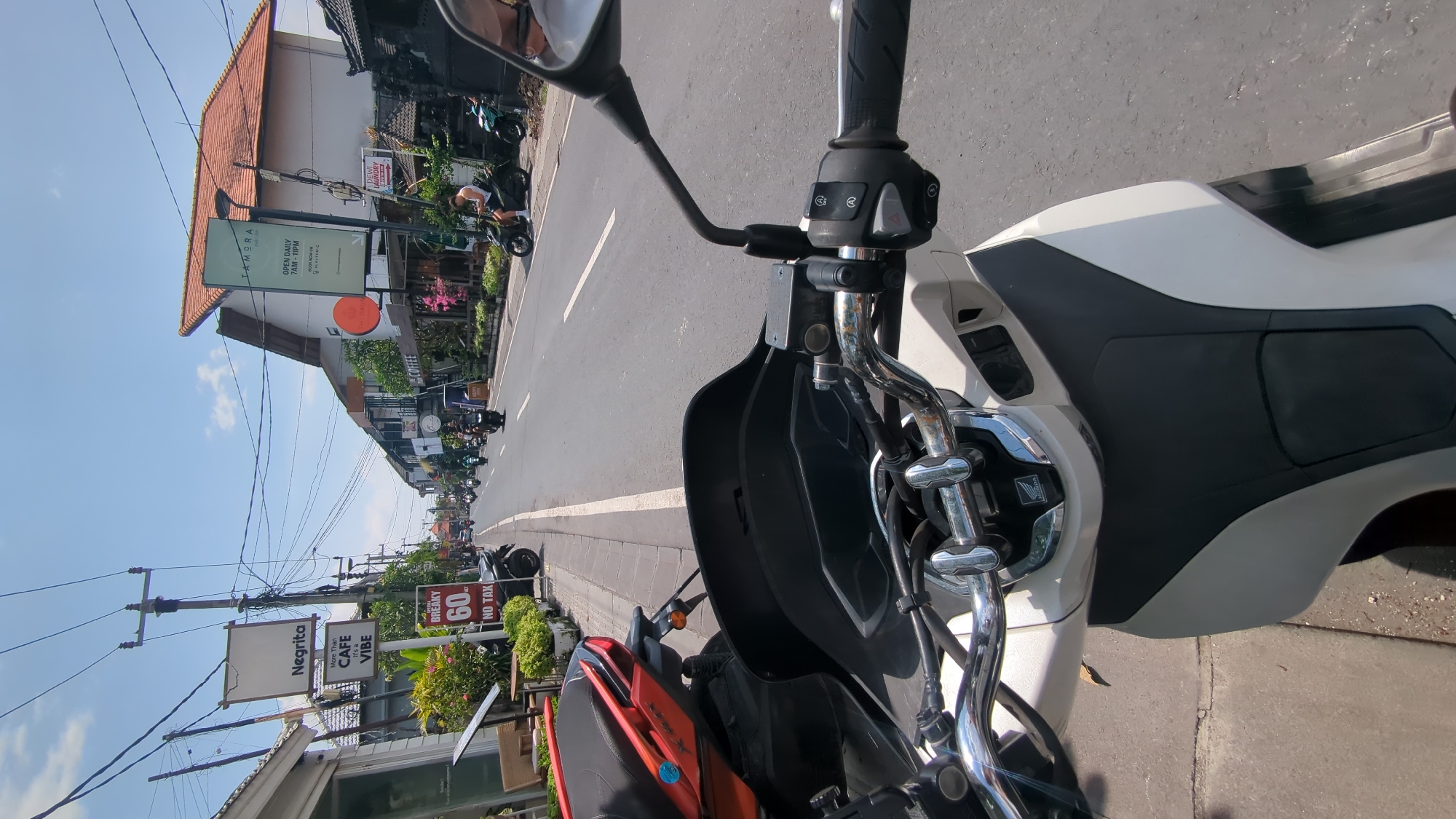 Captured from the perspective of a motorcyclist in North Kuta, Indonesia, the image showcases a street lined with buildings and signs. A Negrita Cafe advertisement is visible, claiming "More Than CAFE, It's a VIBE". Another sign advertises breakfast deals at "60 NO TAX". A sign for "IAMBRA" is displayed with the hours "OPEN DAILY 7AM-11PM" and the "PLAYTOMIC" logo. Street traffic, including motorcycles, extends down the road. The sky is partially visible with a blue sky.