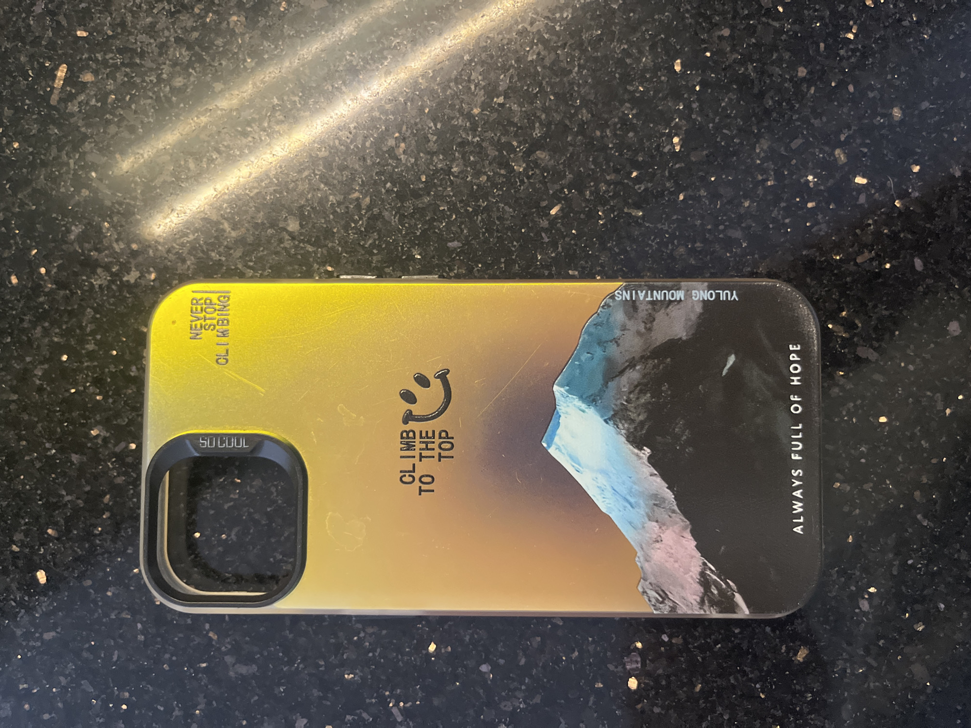 Here is a description of the image:
The image shows a phone case resting on a dark, speckled surface. The case is primarily yellow and black and features a stylized mountain image. The words "SO COOL" are written near the camera lens opening. Other text elements include "CLIMB TO THE TOP" with a smiley face, "NEVER STOP CLIMBING", "ALWAYS FULL OF HOPE", and "YULONG MOUNTAINS". The location in Bac Nha Trang Ward, Vietnam is not discernible from the image.