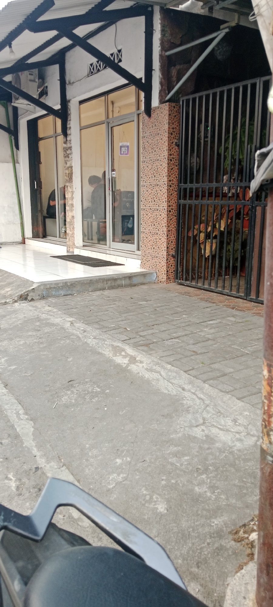 The image depicts the exterior of a commercial establishment in Maguwoharjo, Indonesia. The building features large glass doors and windows, with a prominent "OPEN" sign visible inside one of the windows. A black metal gate with vertical bars is to the right of the entrance, partially obscuring a planter with foliage. The ground in front of the building is paved with concrete and interlocking bricks. In the foreground, the handlebars and part of the seat of a dark-colored motorcycle are visible, indicating the perspective is from a rider. The overall scene suggests a street-level view of a shop or office, likely during daylight hours based on the lighting.
