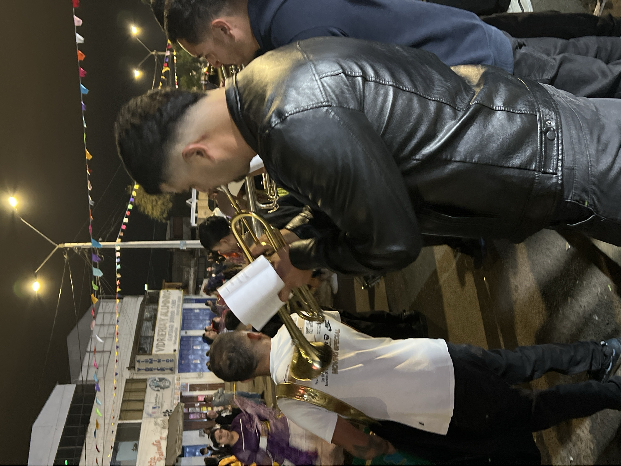 In Arica, Chile, a group of musicians plays brass instruments at night in a street procession. A man in the foreground wears a black leather jacket and plays a trumpet, while another man behind him, wearing a white t-shirt with gold straps, also plays a brass instrument. A woman in a purple dress with feathered embellishments is visible to the left, suggesting a festive event. The background shows buildings with signs for "VIDRIERIA Y ALUMNOS" and "VIVIENDA PAINTS." Colorful triangular flags are strung across the street, illuminated by streetlights. The ground is paved and appears to be a street or plaza. The atmosphere is lively, indicating a public celebration or parade.