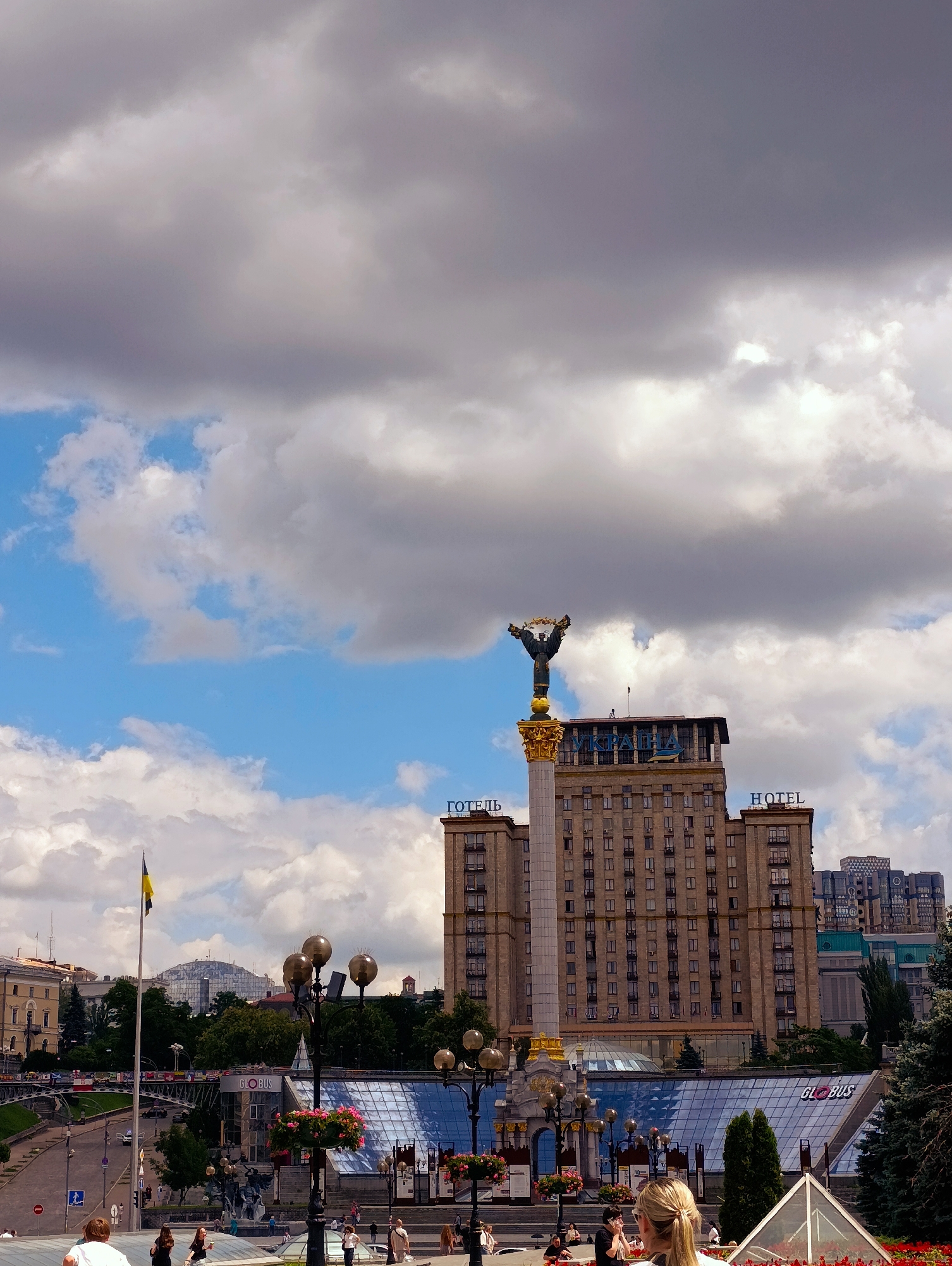 The image depicts a view of Independence Square (Maidan Nezalezhnosti) in Kyiv, Ukraine, on a partly cloudy day. Dominating the center is the Independence Monument, a tall column topped with a statue of Berehynia. Behind the monument, the large, imposing Hotel Ukraina (formerly Hotel Moscow) stands prominently, with "HOTEL" visible in large letters on its facade.
In the foreground, several people are visible, some standing and others seated, suggesting a public space with casual activity. Flower pots with pink flowers are placed along the foreground elements, adding a touch of color. A Ukrainian flag flies on a flagpole to the left of the monument. Further in the background, the "Globus" shopping center is discernible with its glass roof structure. The surrounding architecture suggests an urban environment with a mix of historical and modern buildings. The sky is filled with dramatic, large cumulus clouds against patches of blue.