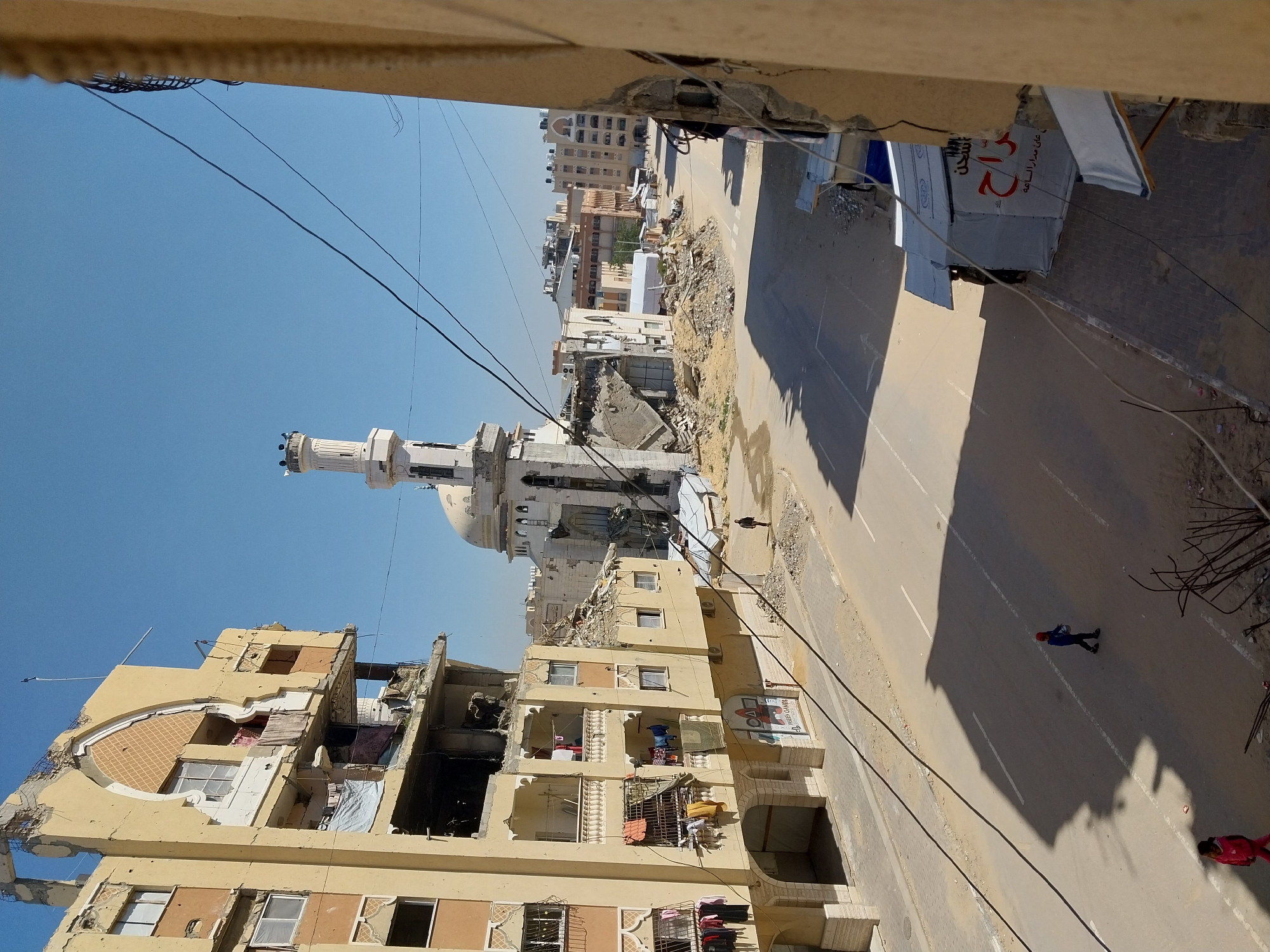 The image depicts a street in a city with significant damage. A mosque with a prominent minaret is visible in the middle ground, alongside buildings that exhibit structural damage, including collapsed sections and debris. Laundry hangs from the balconies of some buildings. The street itself is relatively clear, with two individuals walking along its surface. Power lines stretch across the scene. The sky is clear and blue. The perspective is from an elevated position, likely from a nearby building. The city is possibly located in an urban area of the Middle East.