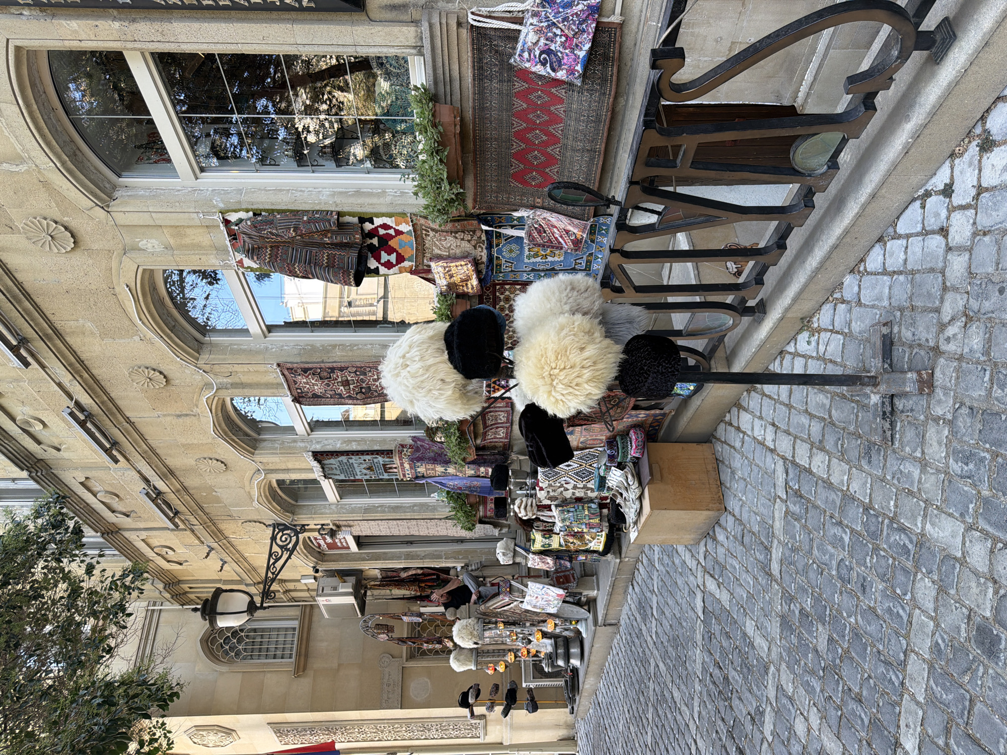 This is an outdoor, eye-level, medium shot of a shop facade on a cobblestone street in the Sabail Raion of Azerbaijan. The building is constructed of light-colored stone with arched windows and decorative carvings above them. The shop displays a variety of merchandise outside, including traditional carpets hung on the wall and racks of fluffy sheepskin hats, as well as bags, and shoes. The street is paved with grey cobblestones. A man is seated on the steps of the shop, gesturing with his hand. The overall impression is of a traditional marketplace.