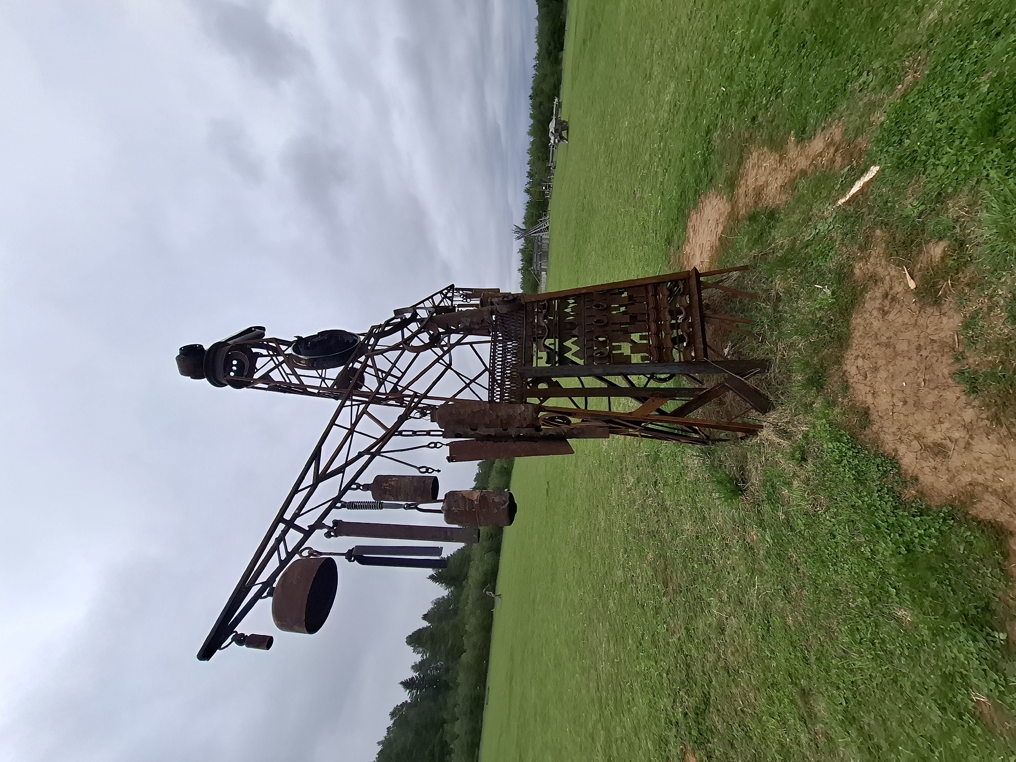 The image shows a large, weathered metal sculpture in a grassy field. The sculpture appears to be constructed from various repurposed metal objects, including pipes, gears, and other industrial components. It has a complex, somewhat abstract design. The field is bordered by trees in the background, and the sky is overcast. The ground around the sculpture's base is bare and appears to be compacted. The overall impression is that of a rural setting with an artistic installation. The location is not explicitly stated to be in the city of Адам, Russia, so this cannot be confirmed from the image alone.