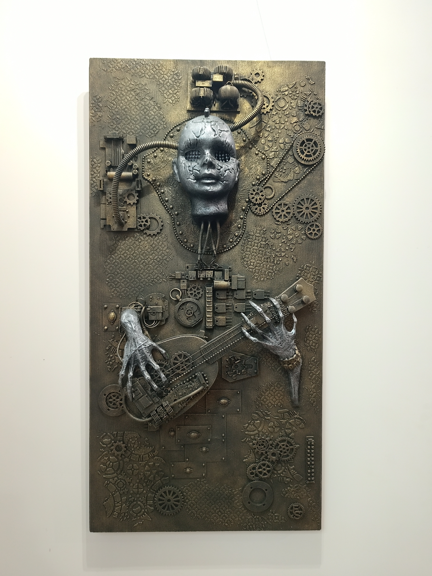 A mixed-media sculpture hangs on a white wall. The piece features a rectangular wooden board painted in metallic bronze and gold tones, with an arrangement of mechanical and organic elements. A silver, mask-like face with hollow eye sockets is positioned at the top, connected to the board by metallic pipes and gears. Below the face, two skeletal hands sculpted in silver grasp a guitar or ukulele. The board is decorated with various mechanical components, including gears, cogs, and small boxes, as well as patterns resembling stylized leaves or flowers. The sculpture is likely indoors and professionally lit, as evidenced by the shadow it casts on the wall. The location of the scene is unknown.
