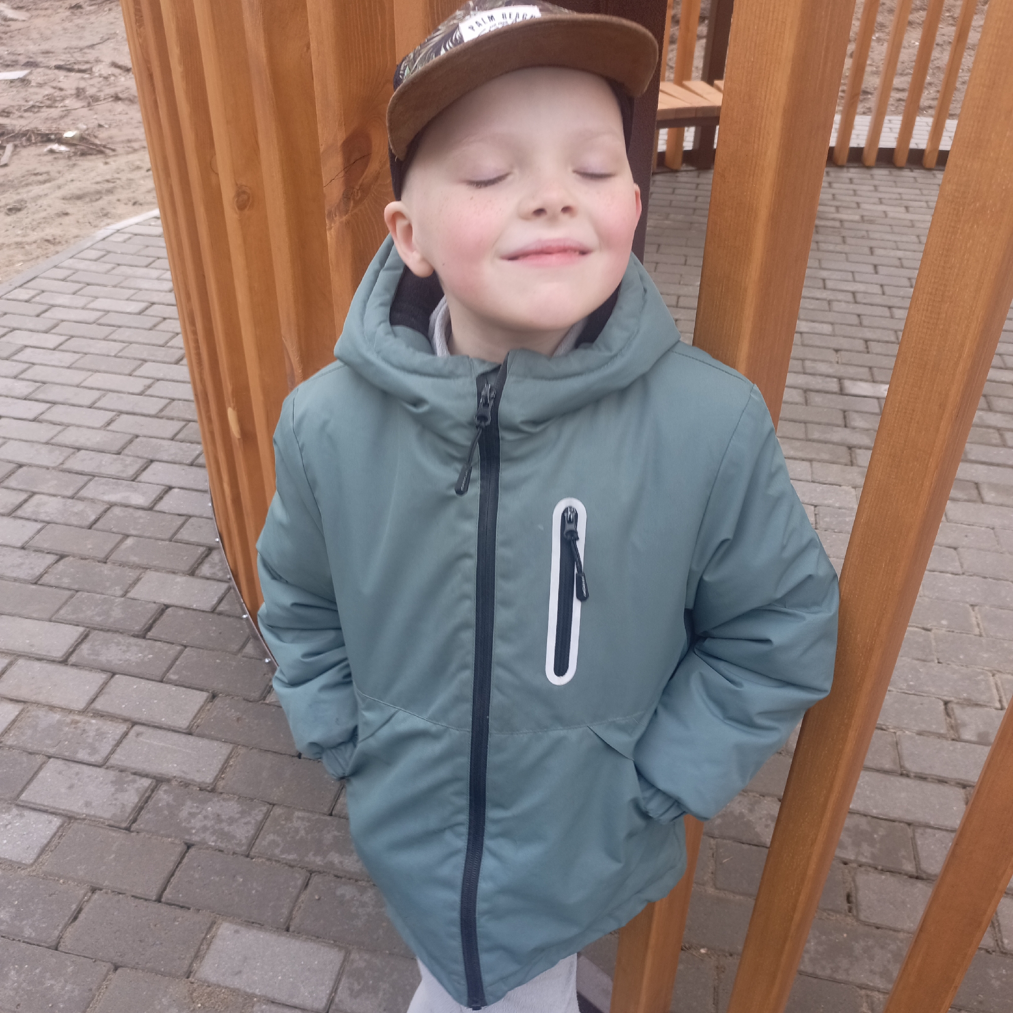 A young boy stands outdoors, centrally framed against a backdrop of wooden slats and paved ground. He has his eyes closed and a gentle smile, suggesting a moment of contentment or enjoyment, possibly basking in sunlight. He is wearing a dark teal or muted green hooded puffer jacket with a black main zipper and a prominent vertical white stripe next to a zippered pocket on the right side of his chest. His hands are casually tucked into his jacket pockets. On his head, he wears a baseball cap with a brown brim, featuring a pattern on the crown, where the words "PALM BEACH" are partially visible.
The ground beneath him and around is covered with rectangular grey paver stones. To his right, and partially behind him, is a curved structure made of light brown wooden slats, resembling a fence, a part of a gazebo, or playground equipment. In the background to the left, there's an expanse of bare, light brown earth or dirt, suggesting an unpaved area. The overall scene conveys a calm, daytime atmosphere, likely in mild or cool weather given the boy's attire.