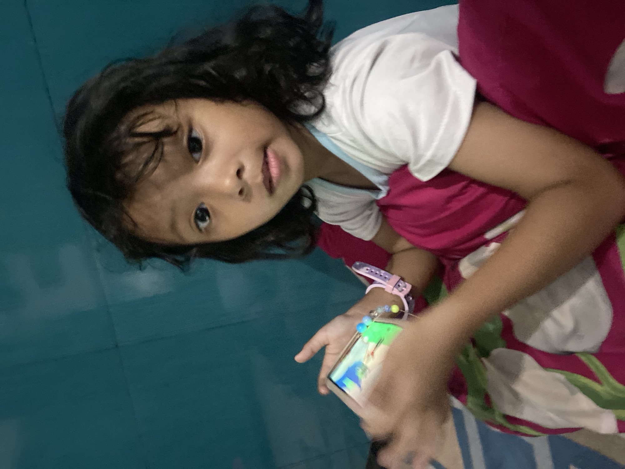 A young girl with dark, curly hair and brown eyes is holding a smartphone in her hands, looking directly at the camera. She is wearing a white t-shirt with a light blue trim and a pink bracelet on her left wrist. She is positioned in front of a teal-colored wall, with a pillow behind her and a floral patterned fabric beneath. The scene appears to be indoors. The image has been taken in Gelam, Indonesia.