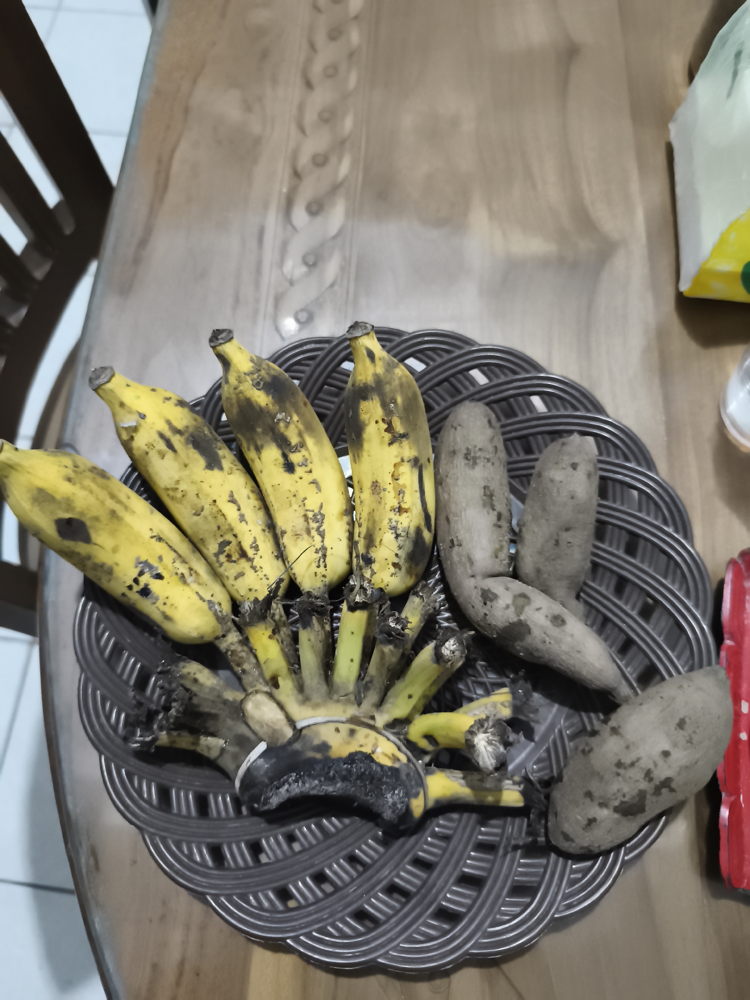 On a wooden table in a home in Yogyakarta, Indonesia, a woven brown basket holds a bunch of yellow bananas with dark spots and three lumpy, brown tubers. The tubers appear to be sweet potatoes or similar root vegetables, still partially covered in soil. The bananas are partially peeled and some show signs of overripeness. The table has a wood grain pattern. In the background, a chair with dark wooden slats is visible, as is a white and yellow package. The scene suggests a simple, domestic setting.