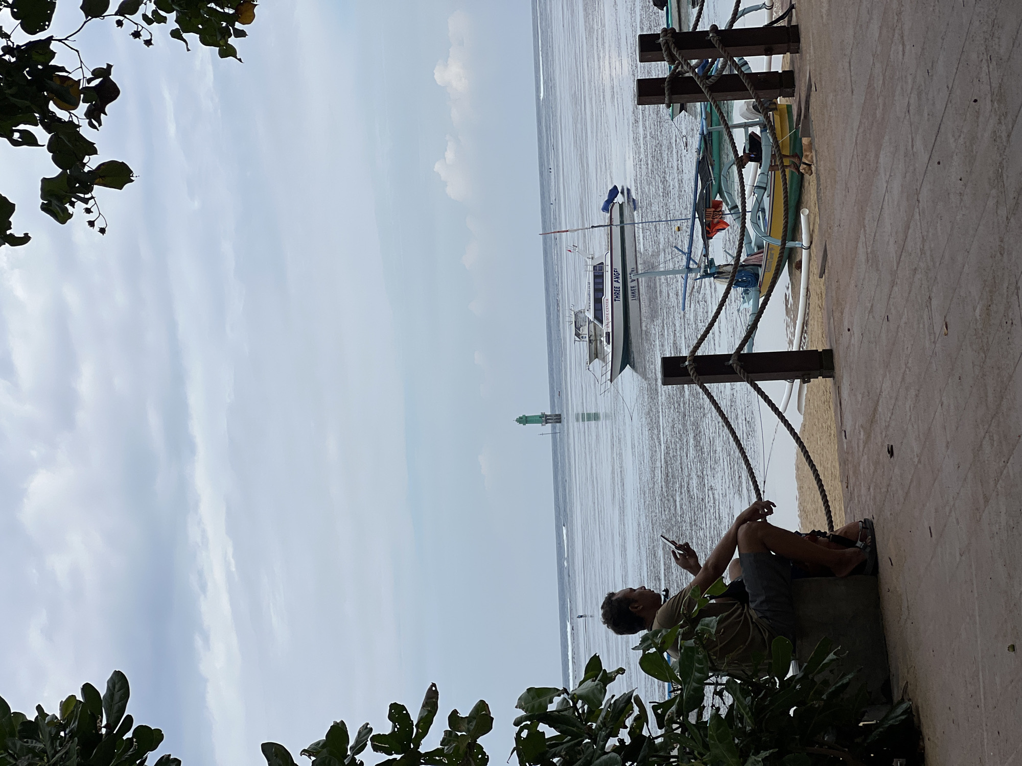 A person sits on a stone bench on a wooden boardwalk, looking at their phone. The boardwalk overlooks a shallow body of water with a sandy bottom. In the distance, several boats are anchored, including a larger white boat with "THREE ANGEL" written on its side and a small green lighthouse. The sky is overcast. The scene is likely a beach or harbor area in Denpasar, Indonesia.