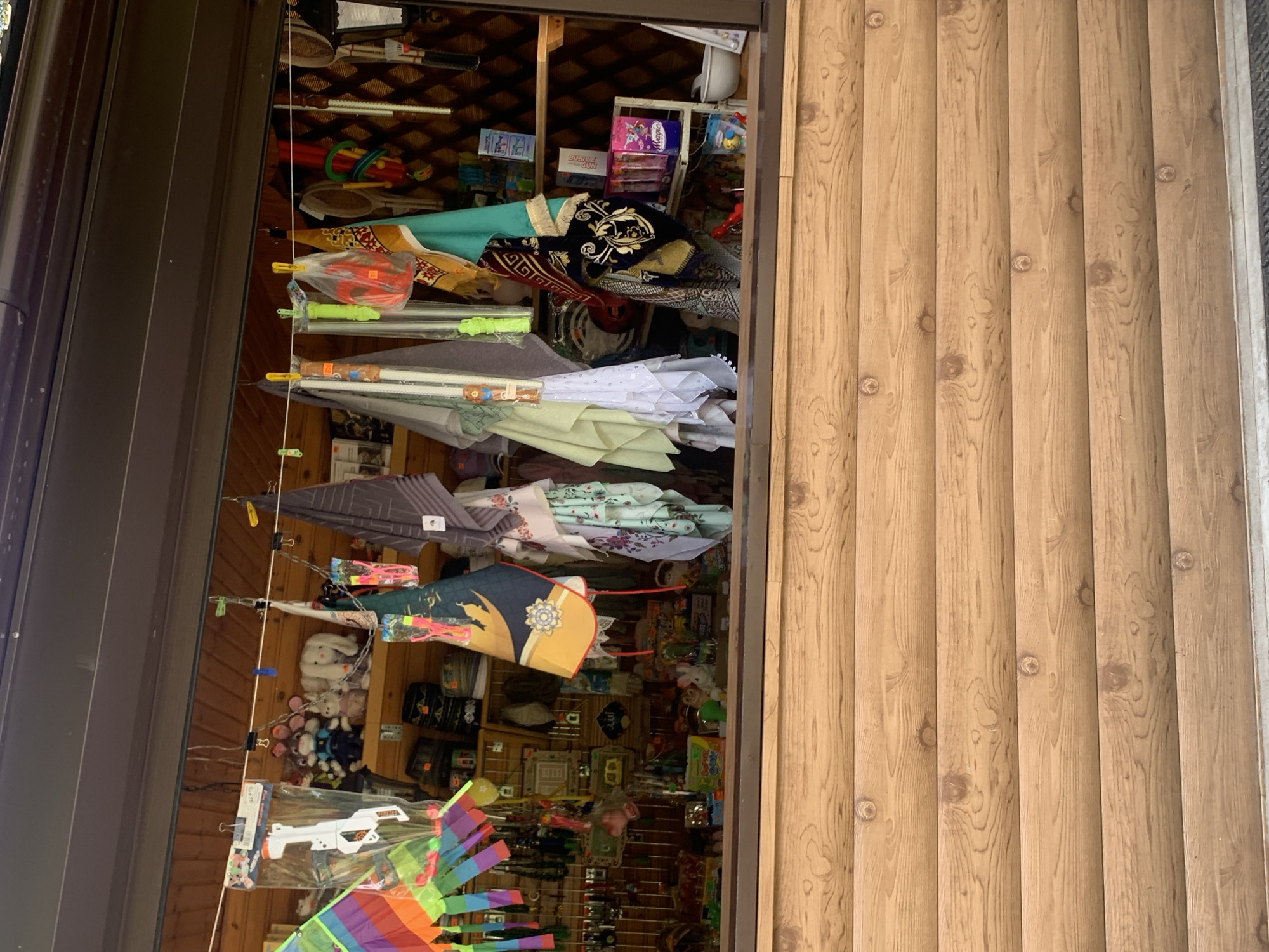 The image depicts an outdoor stall displaying various items for sale, potentially in the city of Arbuzov-Baran, Russia. The stall appears to be a wooden structure with shelves filled with merchandise. Prominently displayed are colorful textiles, plush toys, and assorted novelty items.
In the foreground, a wooden counter or facade with horizontal planks is visible. Behind this, a window or opening reveals the interior of the stall. Hanging from a line across the opening are several items, including a brightly colored kite, various patterned cloths or scarves, and what appears to be a toy gun in plastic packaging. Plush toys, including rabbits and other animals, are arranged on the shelves. Other items visible on the shelves include small boxes, bottles, and other trinkets. The background shows wooden shelving and walls. The overall impression is that of a small shop selling souvenirs or toys. There are no people visible in the image.