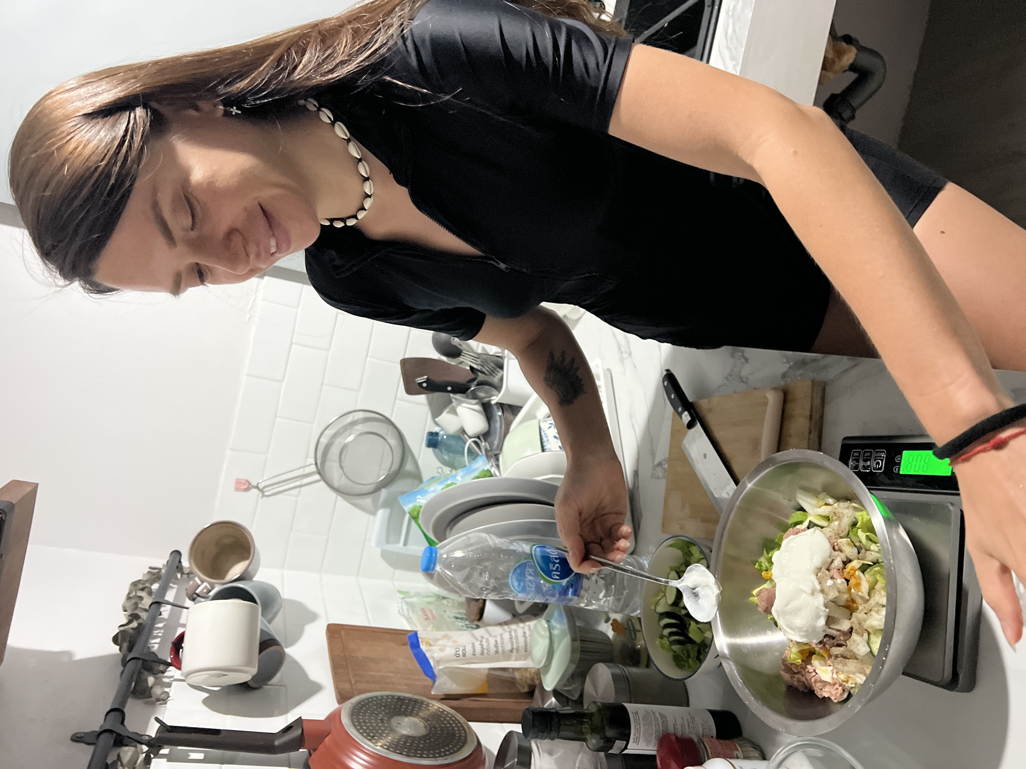 A woman with long brown hair is preparing food in a kitchen. She is wearing a black shirt and a shell necklace. She is adding dressing to a salad in a bowl using a spoon. The salad is on a digital scale, and other kitchen items like plates, bowls, a bottle of water, and cooking utensils are visible on the counter. The backsplash is white tile. The location is in a kitchen in บ้านบางรักษ์, Thailand.