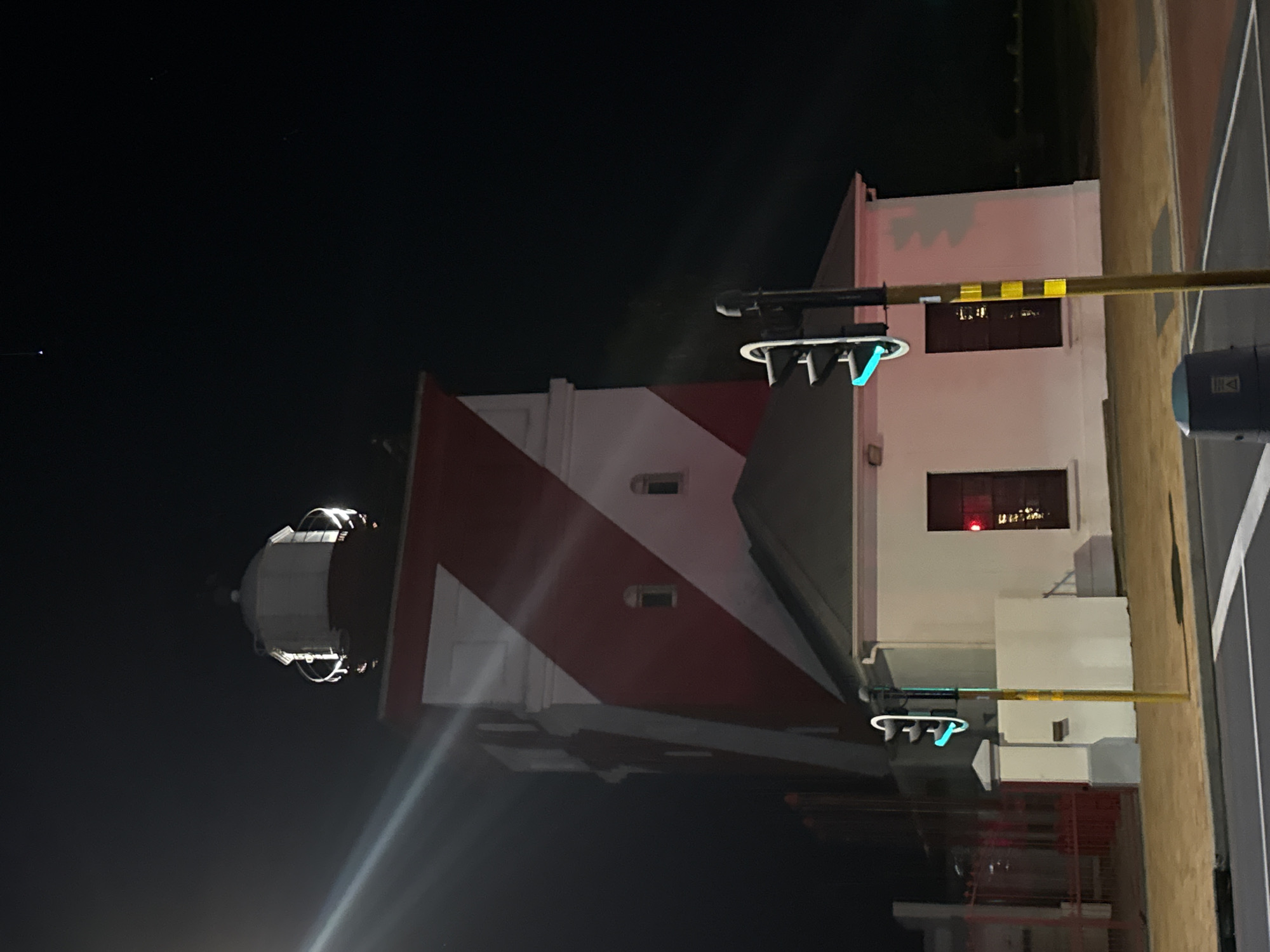 The image depicts the Milnerton Lighthouse in Cape Town, South Africa, at night. The lighthouse is painted with distinctive red and white stripes, standing adjacent to a white building with visible windows. Street lights with green illumination are present, and a portion of the street and sidewalk are visible on the right side of the frame. The background is predominantly dark, representing the night sky.