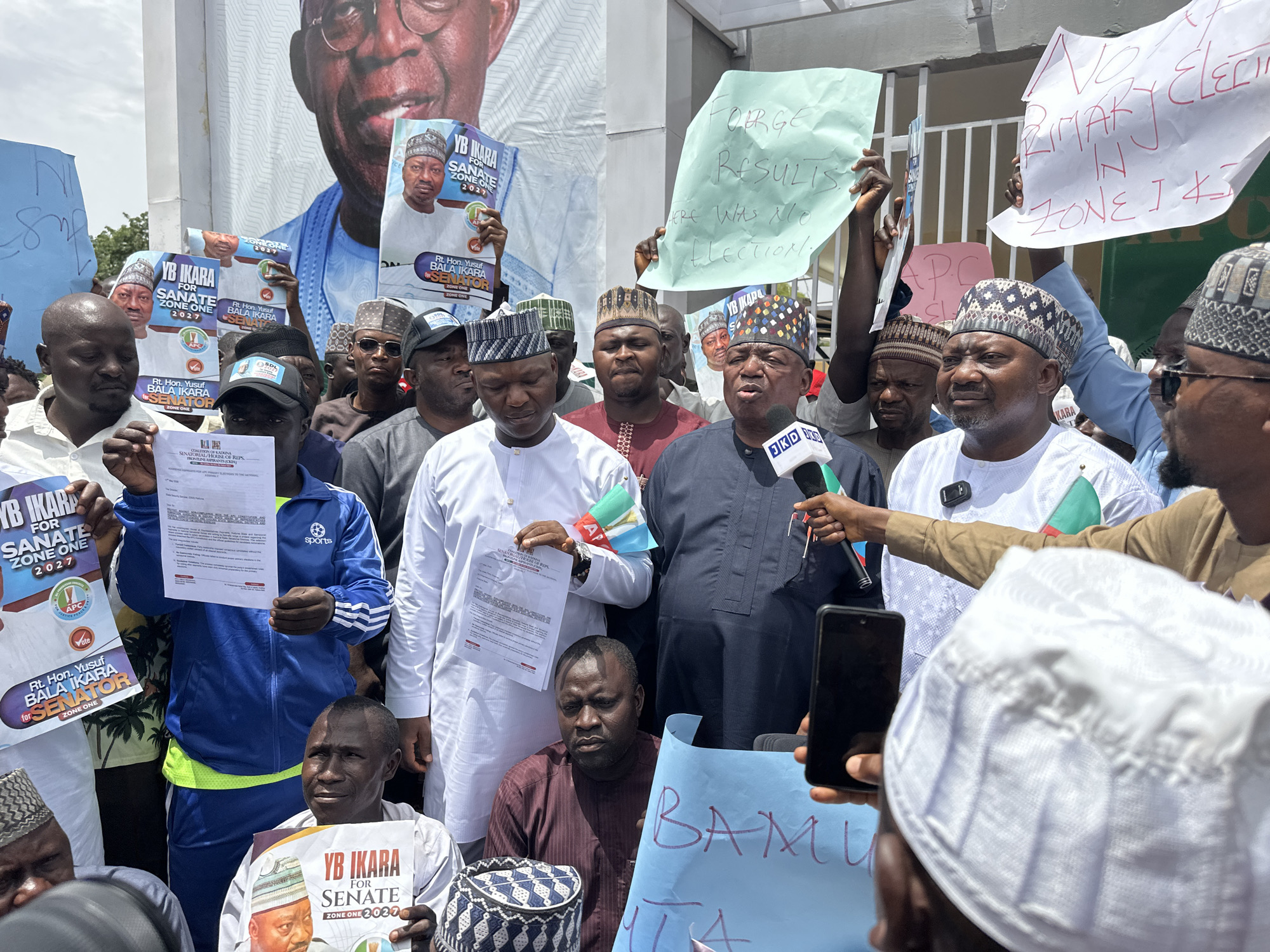 A diverse group of men gathers in what appears to be an outdoor press conference or rally in Maiburiji, Nigeria. Dominating the backdrop is a large poster featuring the face of a man, presumably a political figure.
In the foreground, several men hold signs and posters supporting YB Ikara for Senate Zone One 2027. One sign reads "FORGE RESULTS. HERE WAS NO ELECTION." Another says "No Primary Election in Zone 1 & 3". A man in the center, dressed in traditional attire, speaks into a microphone. A person to his right records him with a smartphone. Another man next to him wears a white outfit.
Attendees sport various hats, including traditional patterned caps and baseball caps. Some hold documents. A Nigerian flag pin is visible on the attire of a man near the microphone. The overall atmosphere suggests a political demonstration or expression of discontent. The environment appears to be urban.