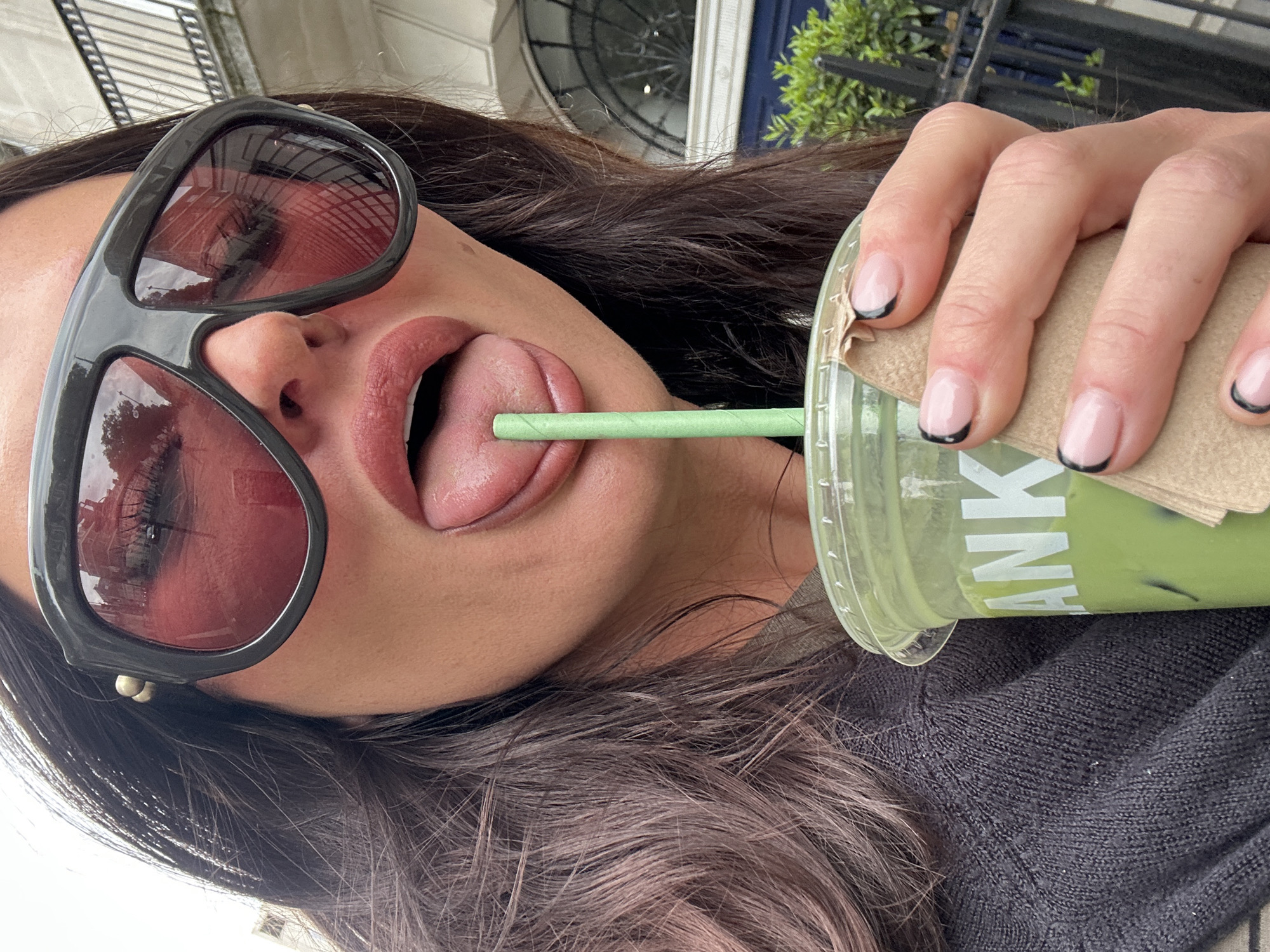 An outdoor selfie taken in daylight features a woman wearing sunglasses, with her tongue extended, and sipping from a plastic cup of matcha green tea with a green straw. The cup's label has "THANK" visible. She is wearing a dark sweater. Her fingernails are manicured with a nude base and black tips. The sunglasses reflect a building façade. In the background are building details and foliage, suggesting an urban environment, possibly the city of London, United Kingdom.