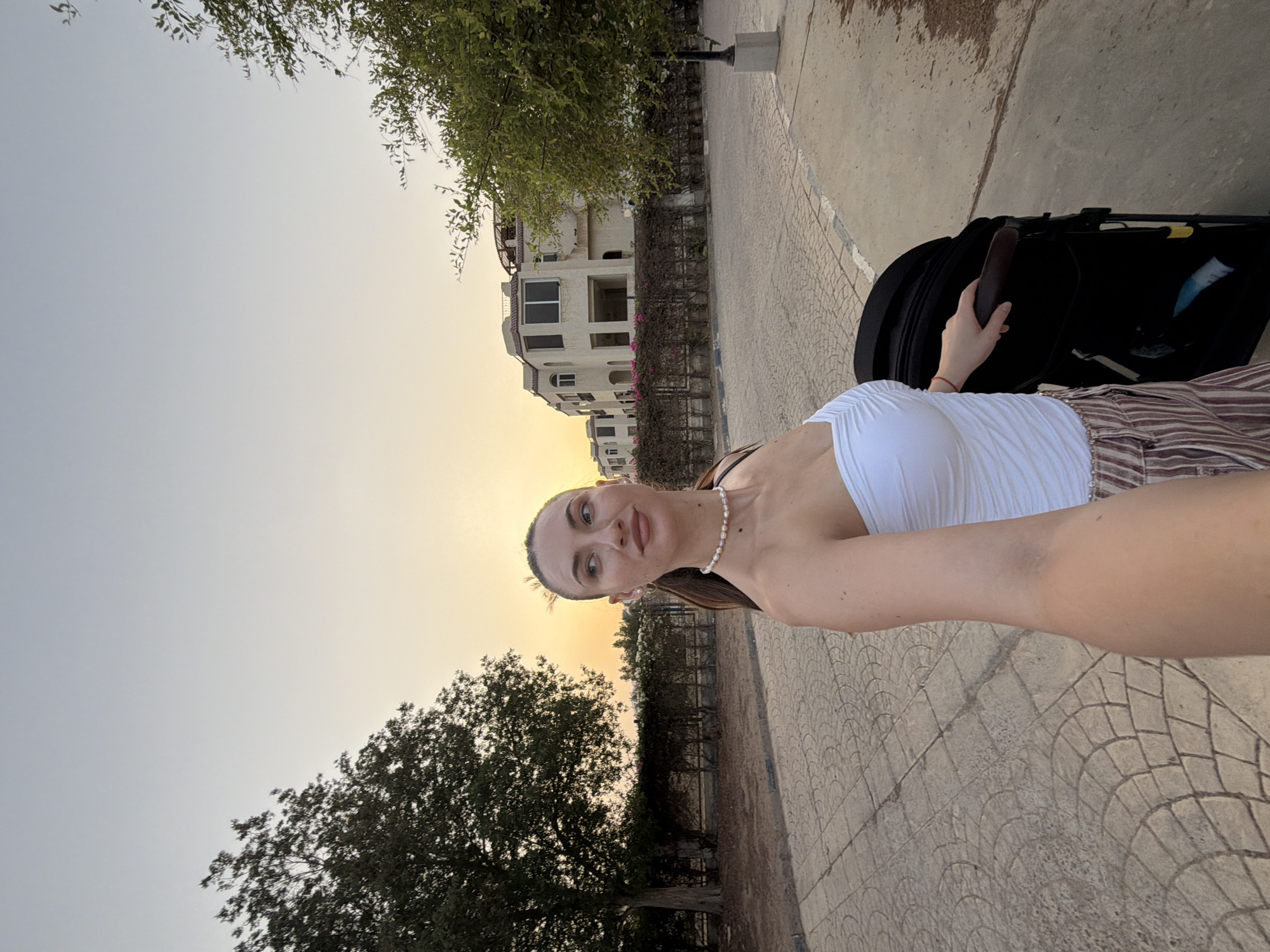 A woman with long, dark hair pulled back in a ponytail, wearing a white strapless top and striped pants, stands on a paved walkway in Sharm El Sheikh, Egypt. She is holding the handle of a black stroller with her right hand and looking to her left. Behind her, several modern buildings with light-colored facades and balconies are visible, situated behind a line of manicured hedges. The sky is a soft gradient of pale blue and orange, indicating either sunrise or sunset. To the left of the woman, a large tree with dense foliage stands beside the walkway, and to the right, another smaller tree is partially visible. The walkway is paved with decorative stone tiles.