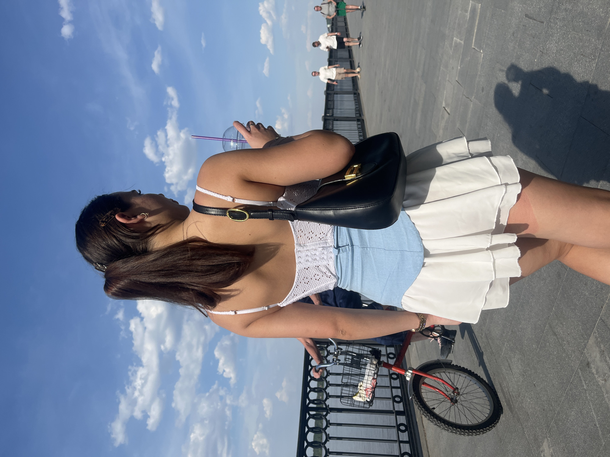A young woman with dark hair styled in a low ponytail stands on a paved walkway in Perm, Russia, near a black metal fence overlooking a body of water. She is holding a clear plastic cup with a purple straw. She wears a white top, a light blue denim skirt, and a white ruffled overskirt. A black purse with a gold clasp is slung over her shoulder. She is standing beside a red bicycle with a wire basket on the front. Several figures are walking in the background on the same walkway. The sky is blue with scattered white clouds. The time of day appears to be mid-afternoon based on the shadows.