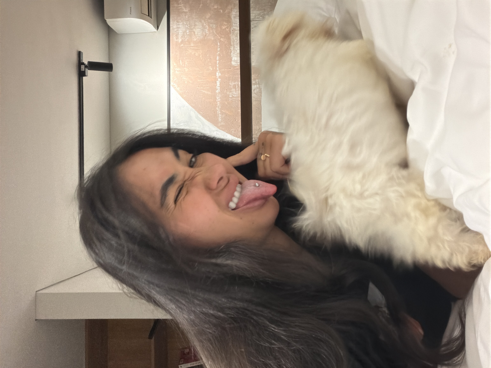 A sideways indoor shot in Tibubeneng, Indonesia, features a woman with long, dark hair and a tongue piercing making a face. She’s touching her face with a hand that has a ring on one finger. A fluffy, light-colored dog is near her face, partially obscured by white bedding. In the background are neutral-toned walls, a wooden panel, and a wall-mounted air conditioner unit with an attached black object. A shelf is also visible in the background.