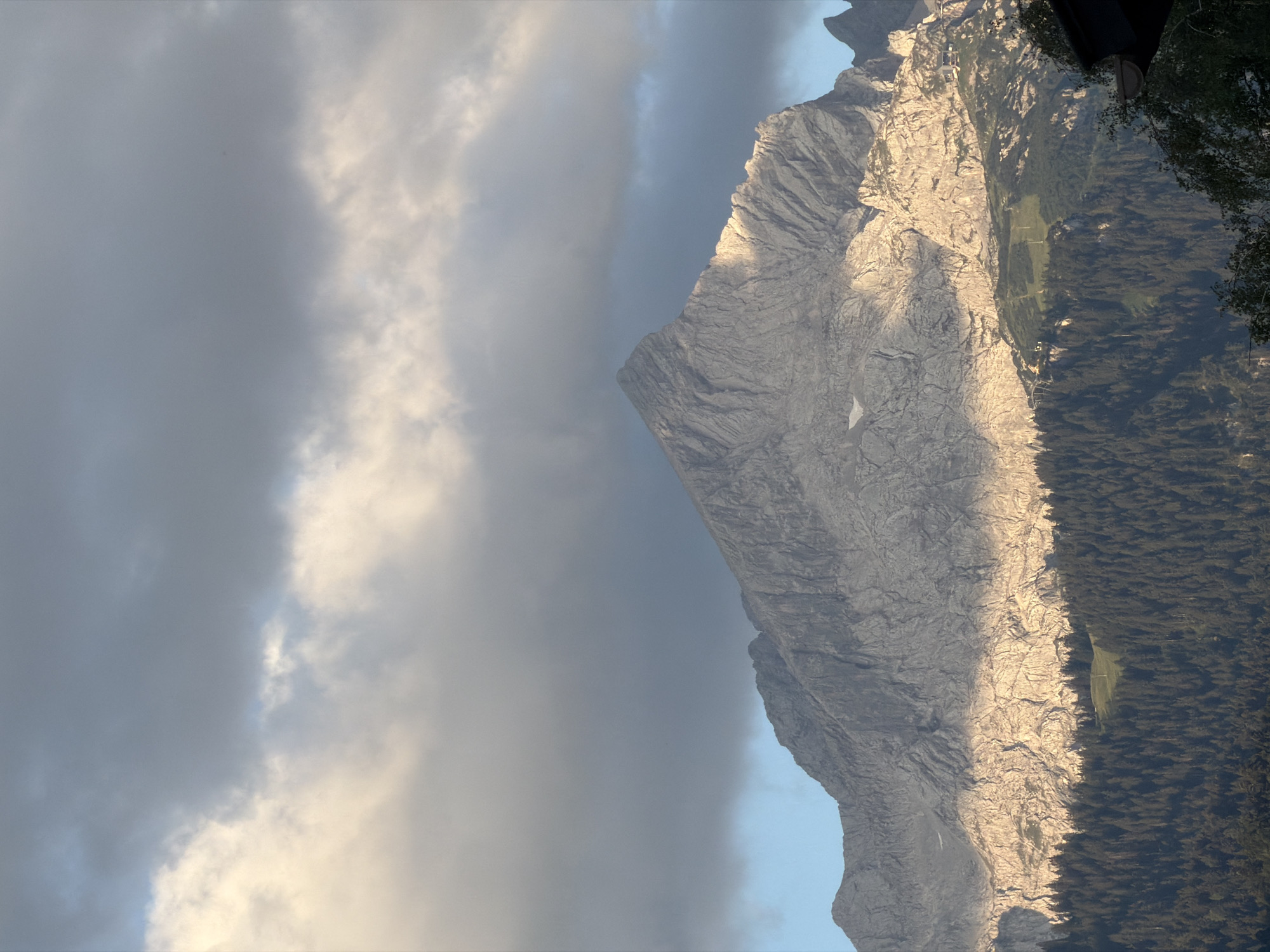 The image depicts a majestic mountain range with rugged, rocky peaks under a dramatic sky filled with dark, heavy clouds interspersed with lighter, illuminated patches. The sunlight casts a warm glow on the lower slopes of the mountains, highlighting textures and creating a stark contrast with the shadowed upper regions and the dark forest that blankets the base. A section of a dense, dark green forest occupies the lower portion of the frame, with a small, isolated patch of bright green grass visible within it. The scene suggests a natural, outdoor environment, likely in a mountainous region. While the image is a landscape and does not contain people or specific urban elements, it evokes a sense of the natural beauty of alpine regions. The context of Garmisch-Partenkirchen, Germany, known for its Alpine landscape and ski resorts, suggests this could be a view of the Wetterstein or Karwendel mountain ranges.