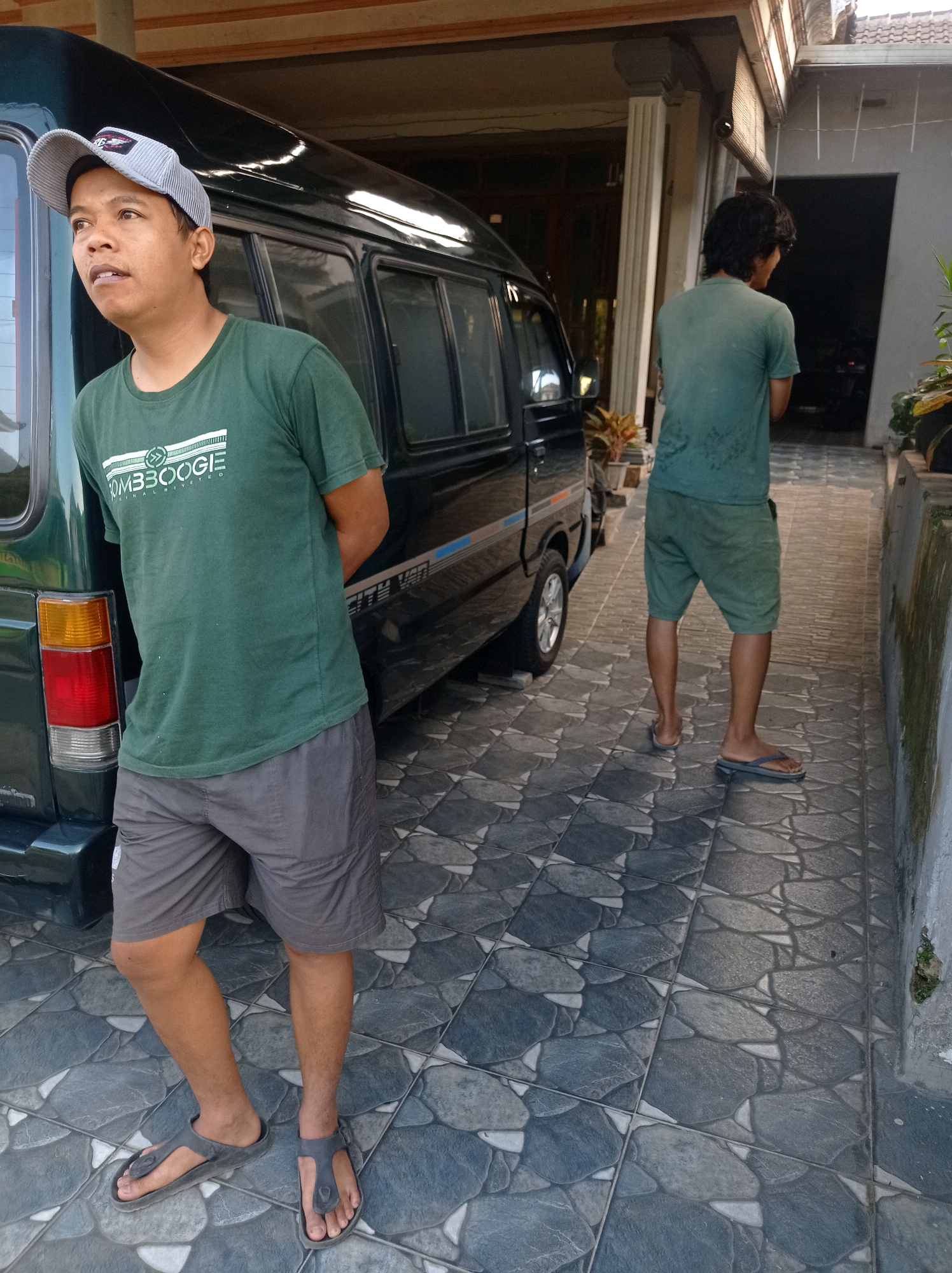 In the city of Purwosekar, Indonesia, two men are captured in a photograph. The man in the foreground, wearing a grey and white striped cap, a green t-shirt with white text, and grey shorts, stands with his hands behind his back. He looks to his left with an expectant expression. To the right and slightly behind him, another man, viewed from the back, wears a faded green t-shirt and green shorts. He is walking away from the camera, looking down. They are positioned on a tiled driveway, with a dark green van parked to the left of the man in the foreground. The background reveals a covered outdoor area and a building with a tiled roof.